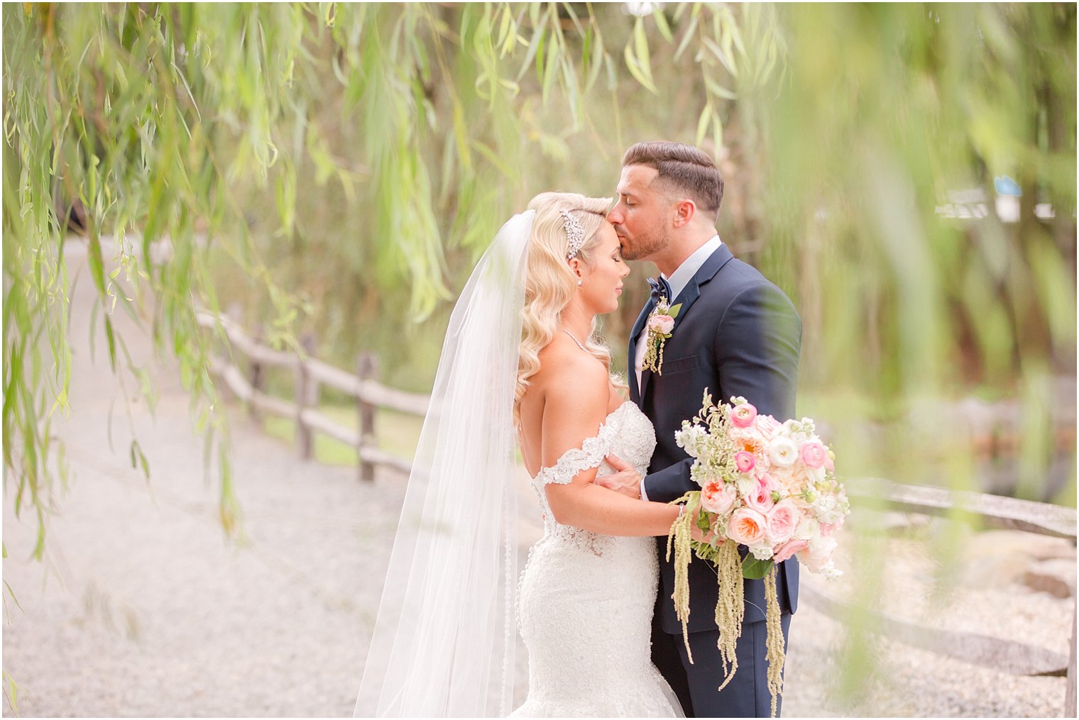 groom kisses bride on forehead in weeping willow at Windows on the Water at Frogbridge