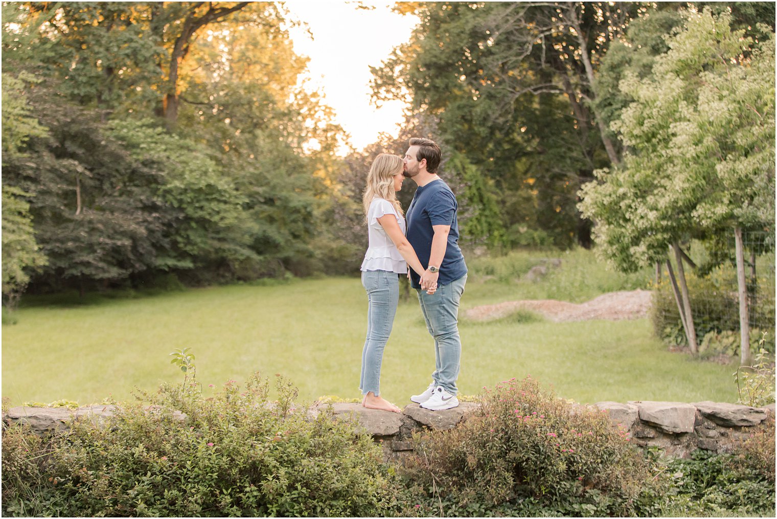 engaged couple holds hands standing on wall at Cross Estate Gardens while groom kisses bride's head