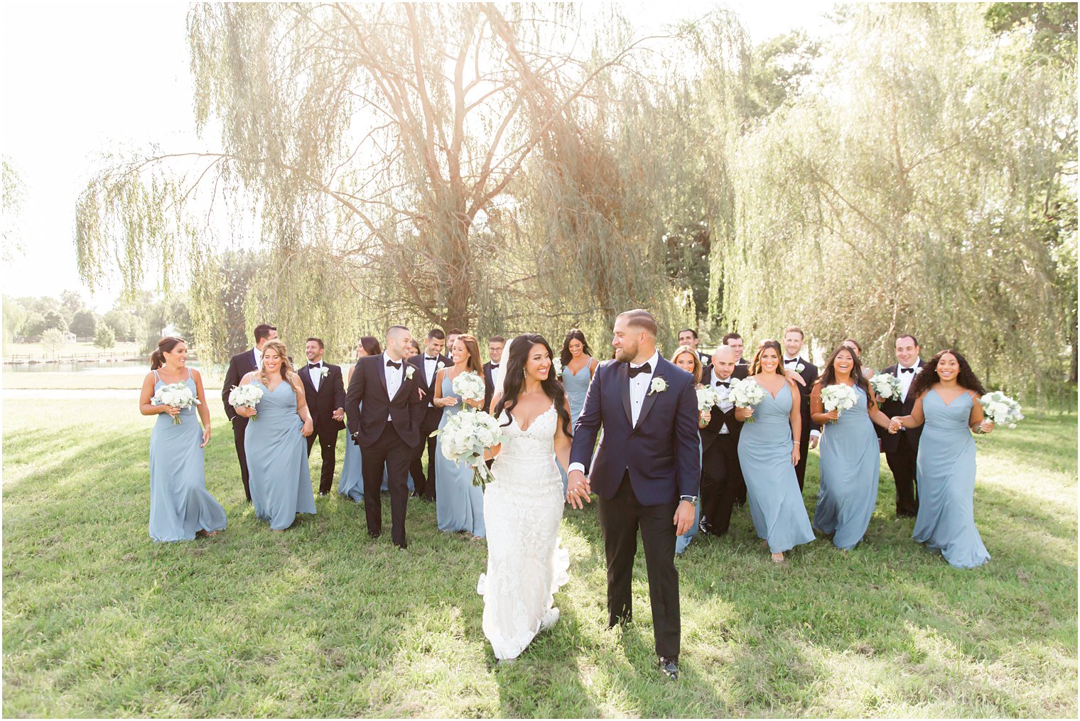 bridal party in black tuxes and blue gowns walk along grass at Windows on the Water at Frogbridge