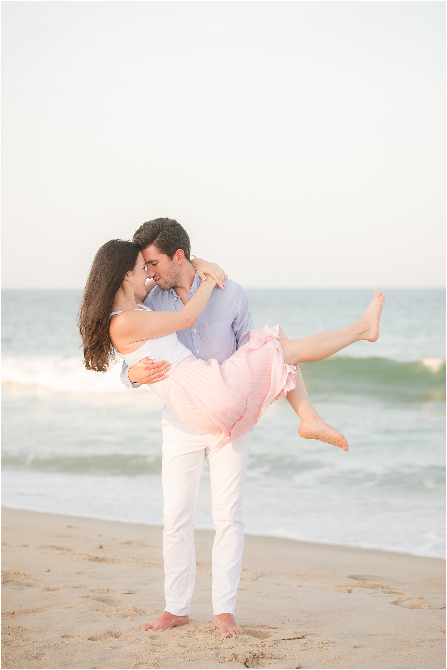 groom lifts bride in pink skirt during beach engagement session
