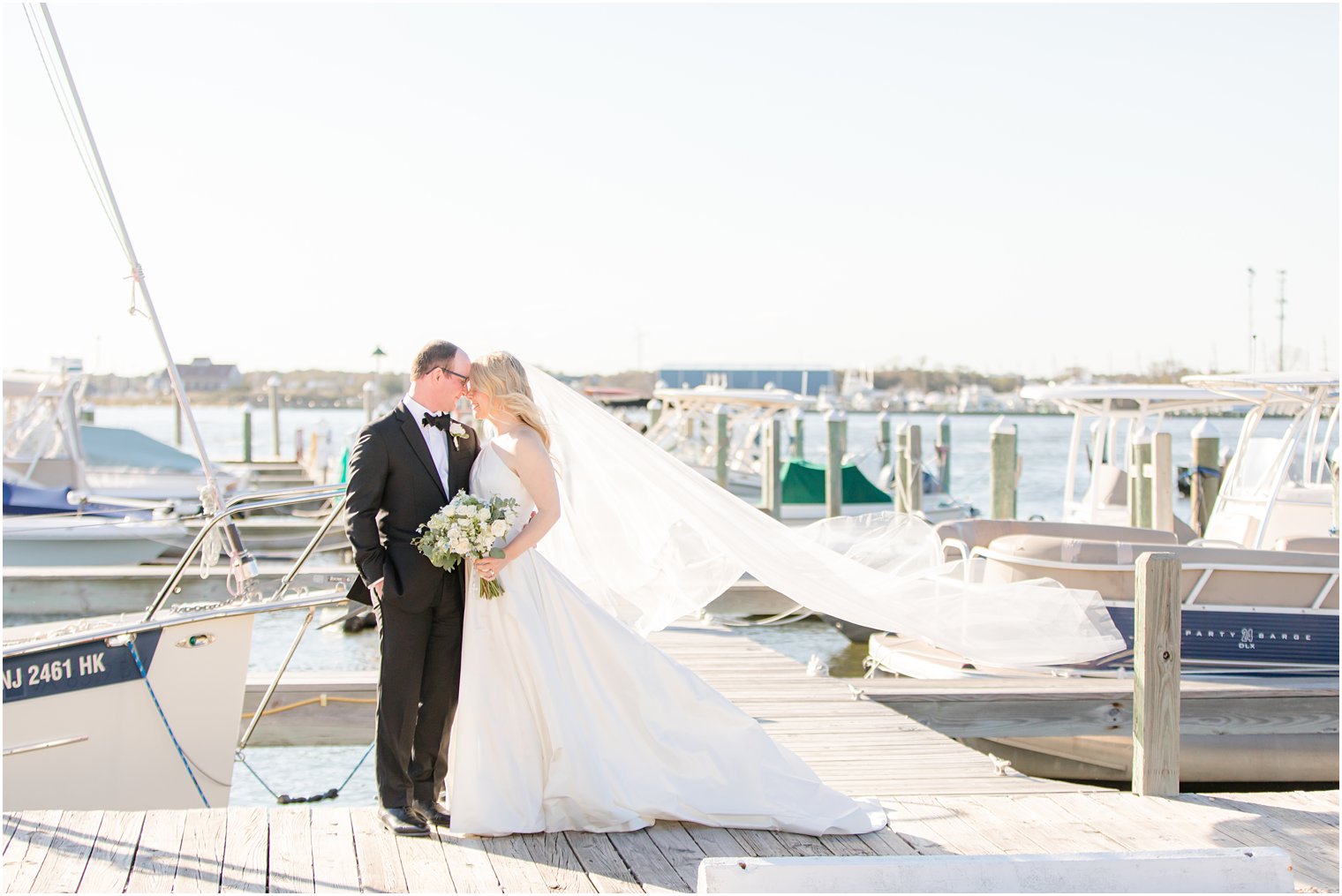 bride and groom pose on pier with bride's veil floating