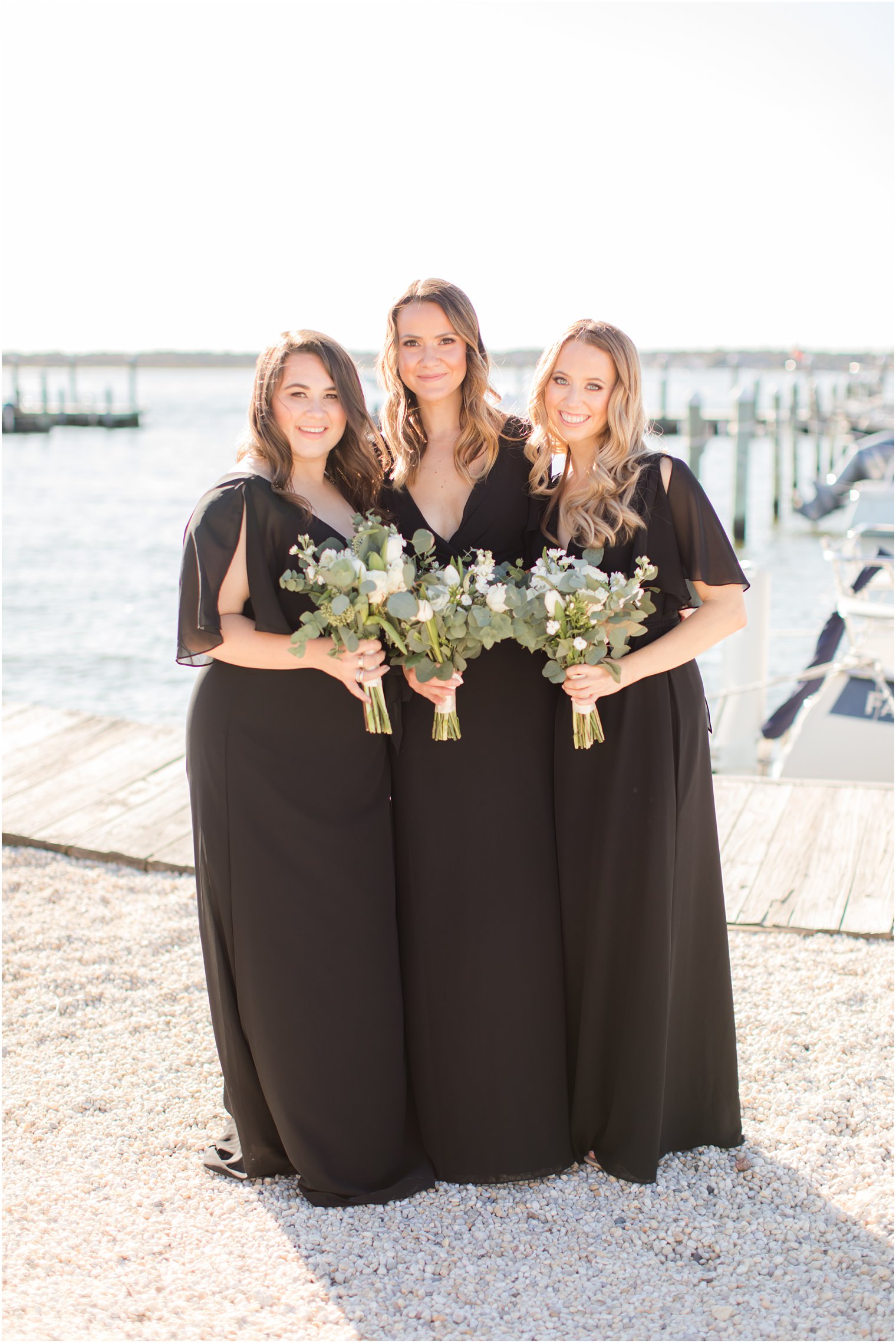 bridesmaids in black dresses hold bouquets with white flowers and eucalyptus leaves