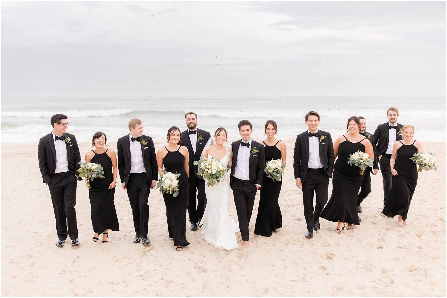 bride and groom walk with bridesmaids in black gowns and groomsmen in tuxes on Spring Lake Beach
