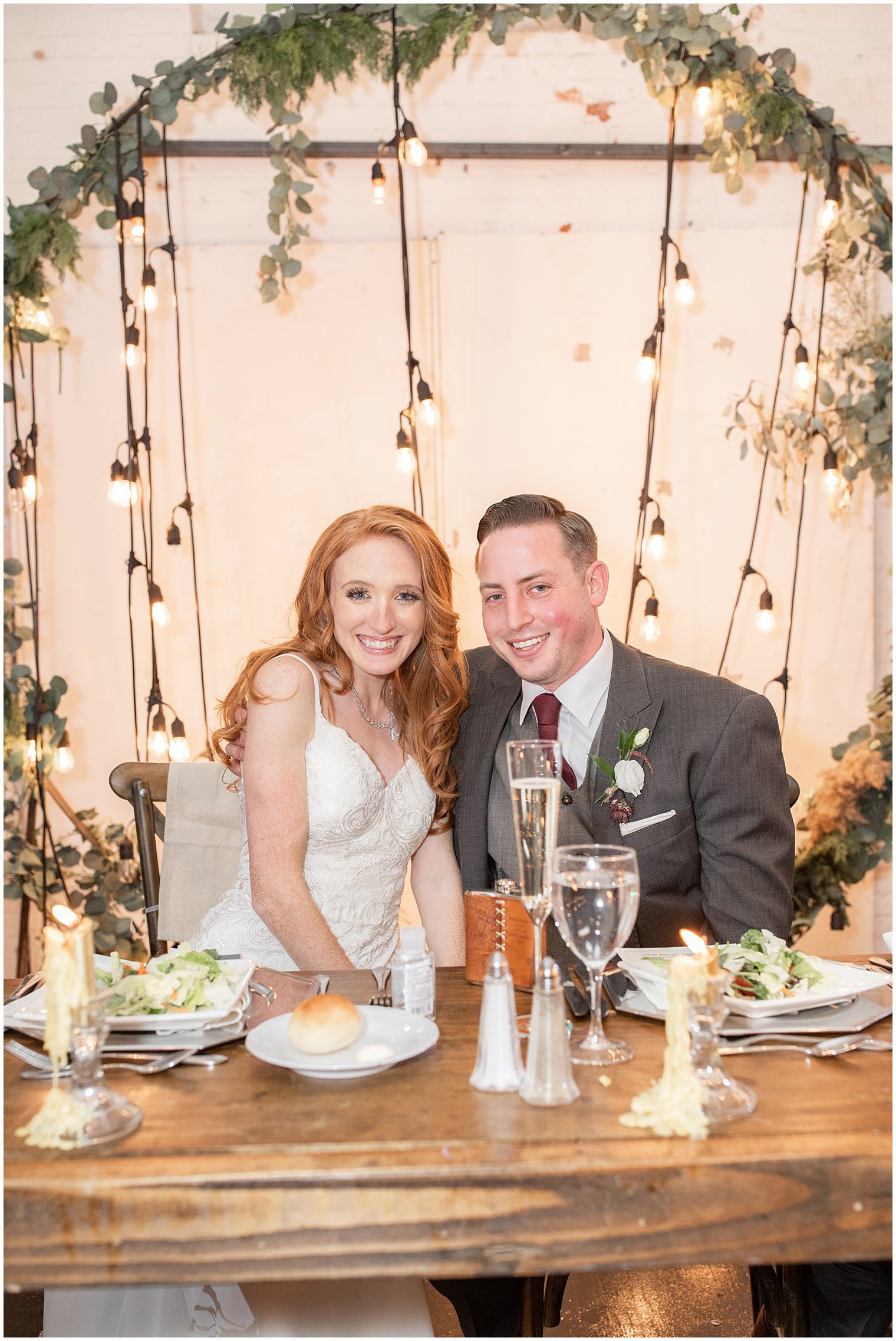 bride and groom sit at sweetheart table with greenery covered metal arbor with lights hanging down behind them
