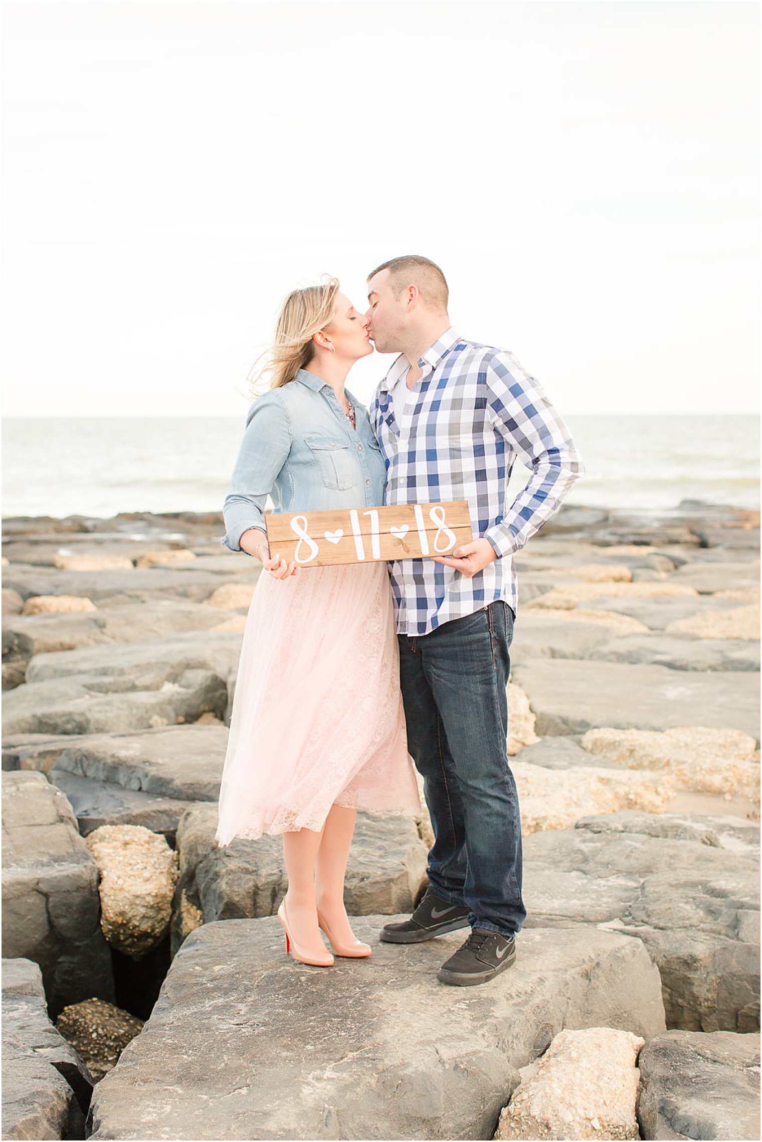 engaged couple stands on rocks at beach holding wooden sign