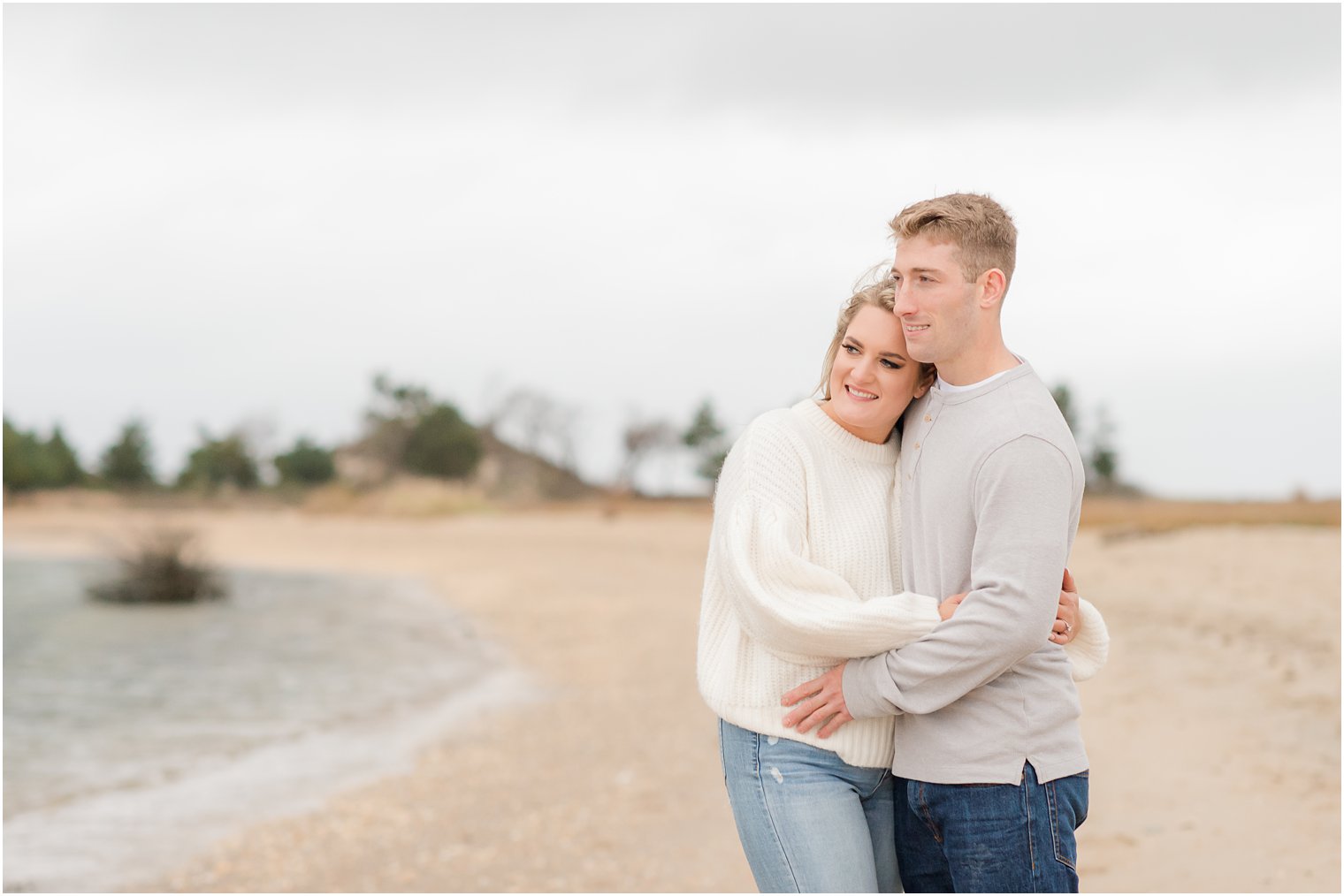 engaged couple hugs on beach looking over water in Sandy Hook NJ