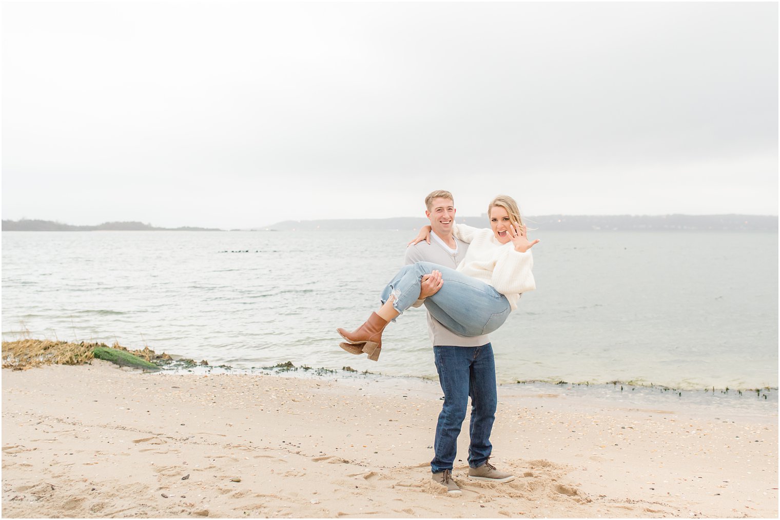 groom holds bride on beach while she shows off engagement ring