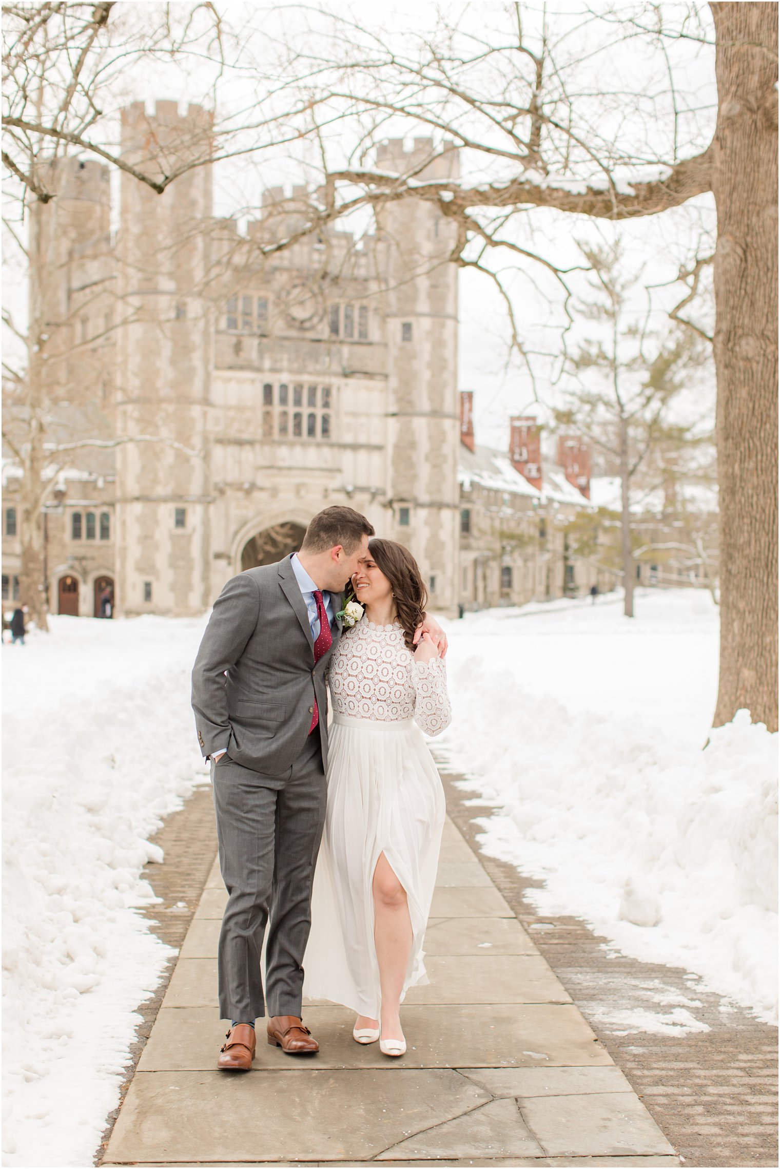 bride and groom walk along sidewalk at Princeton University in the snow leaning down to kiss 