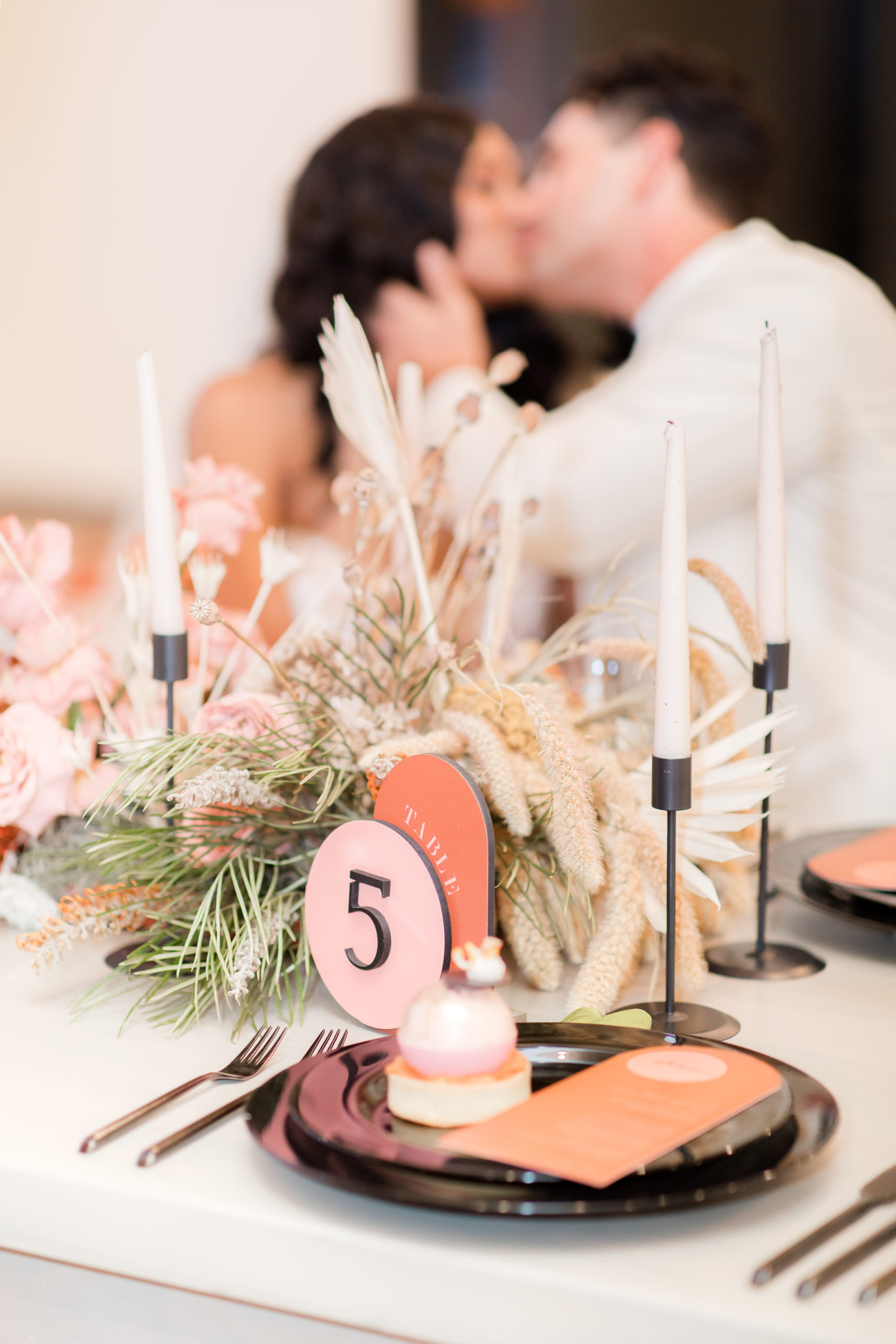 bride and groom kiss in background while wedding dessert sits on black plate during Ironworks at Pencoyd Landing wedding styled shoot