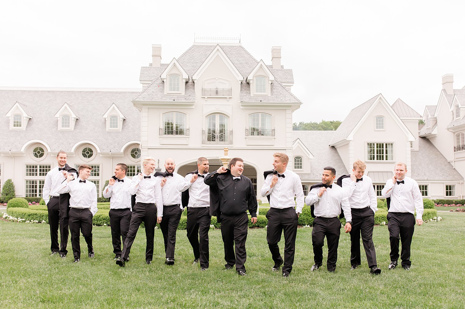 groom and groomsmen walk on lawn at Park Chateau Estate with jackets over shoulders 