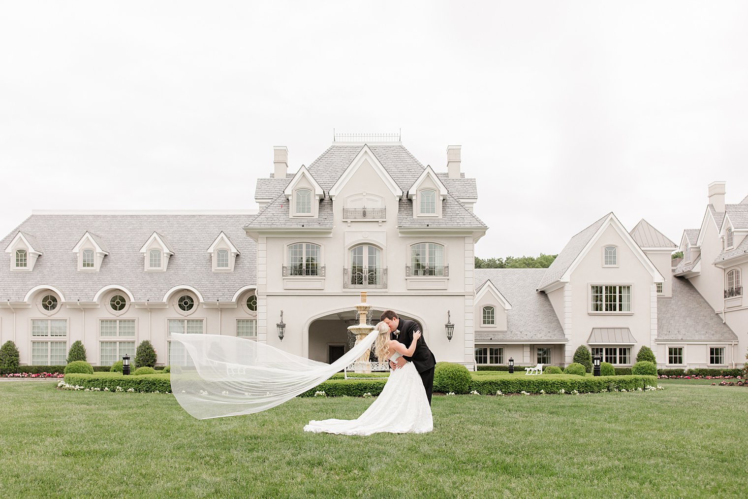 newlyweds kiss on lawn at Park Chateau Estate with bride's veil floating behind her