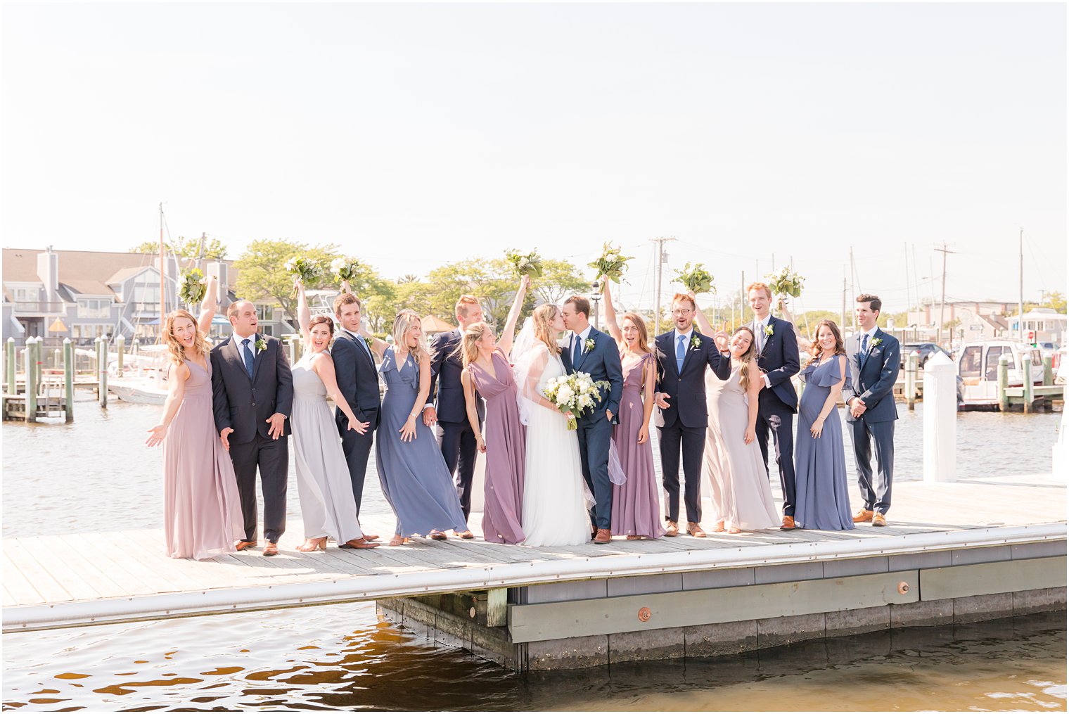 bridal party cheers standing on pier while bride and groom kiss 