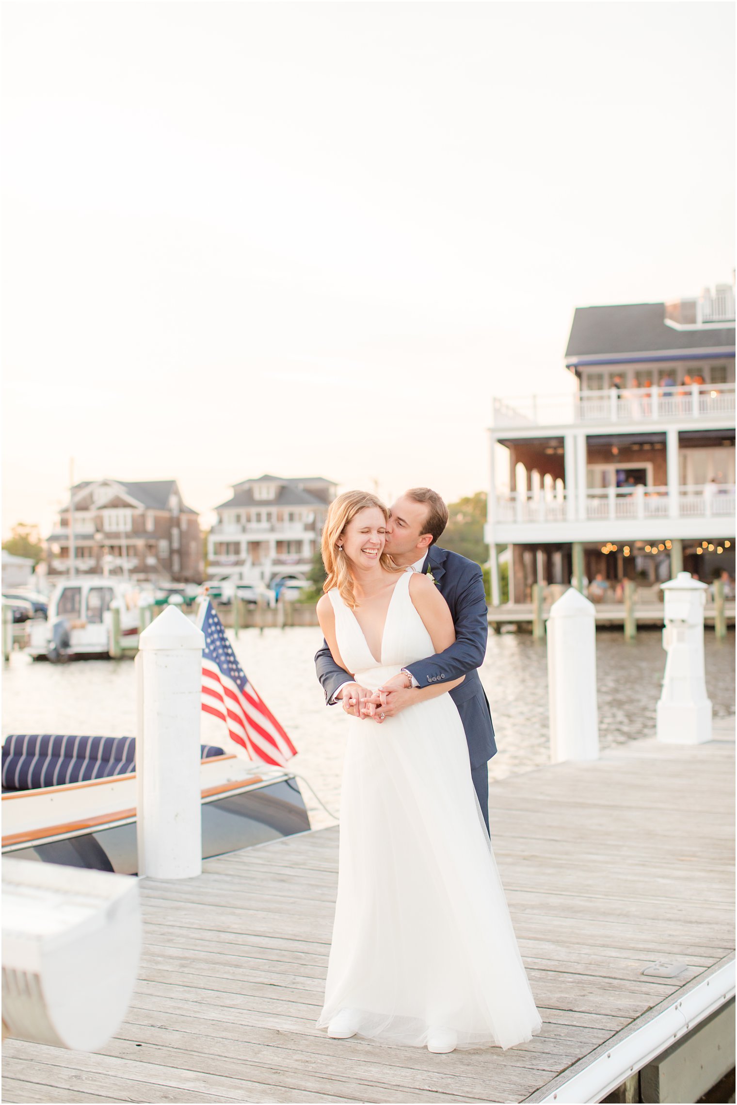 groom kisses bride on cheek while hugging her from behind standing on pier at Bay Head Yacht Club