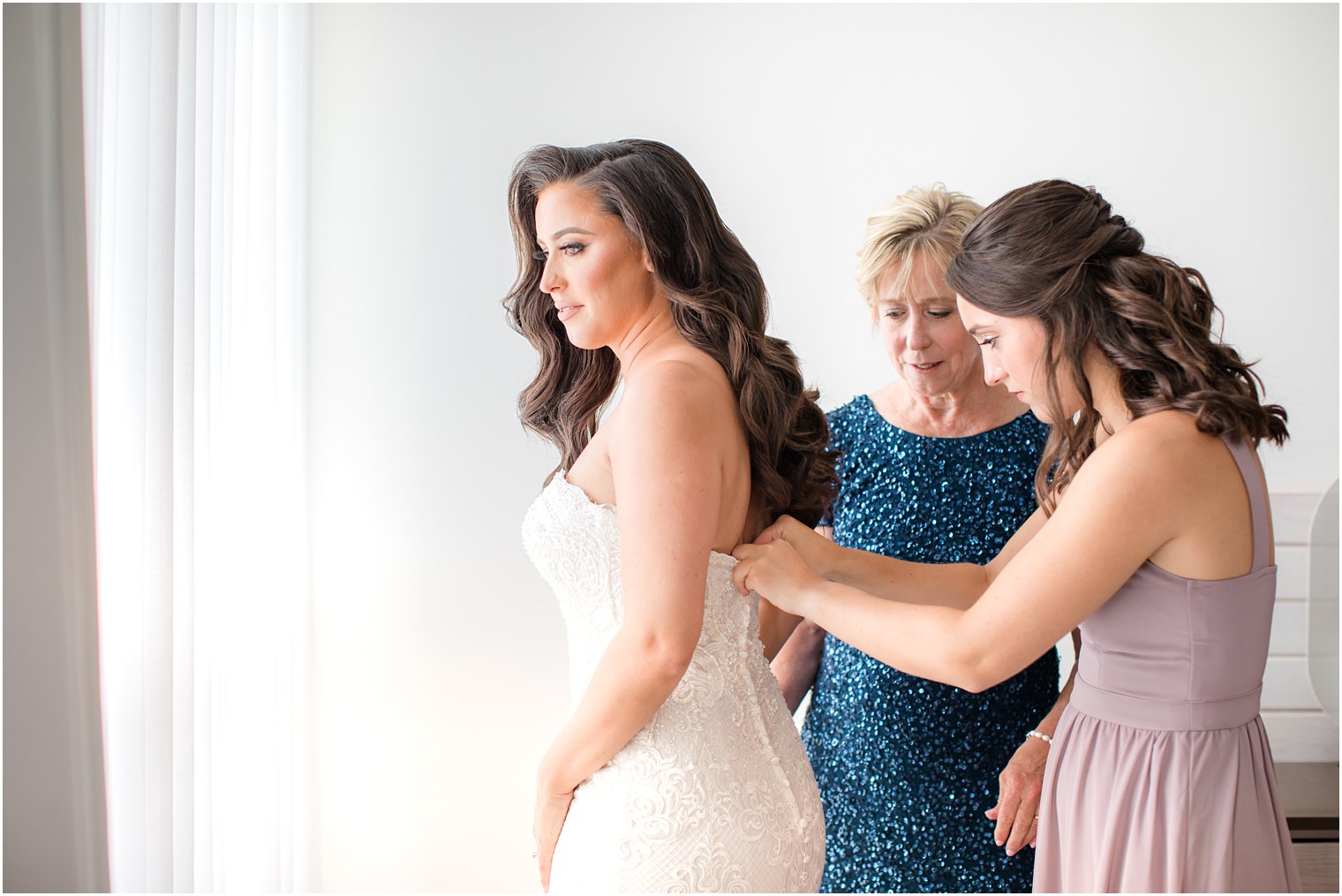 bridesmaid and mother of bride help her into wedding gown