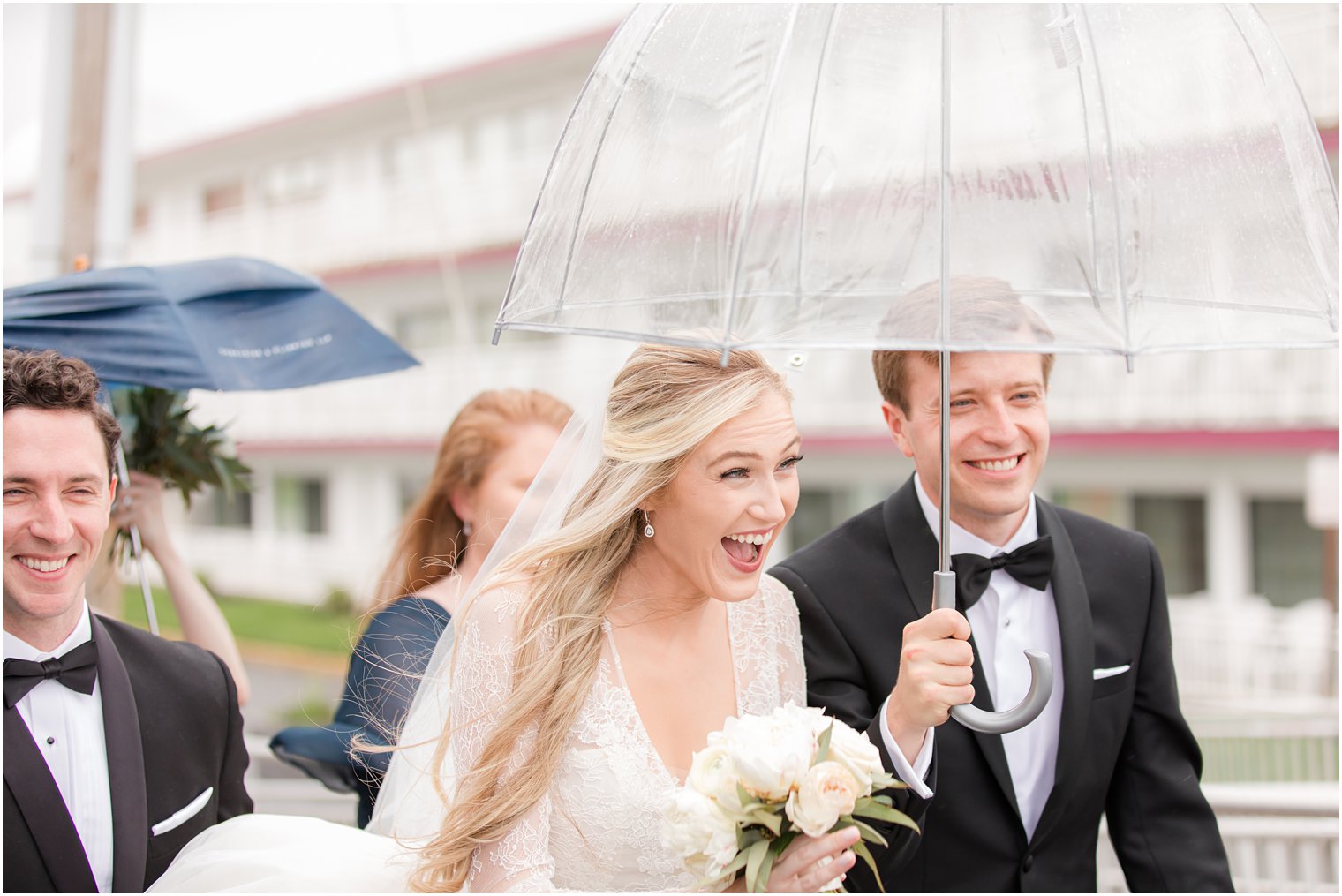 bride and groom laugh under clear umbrella walking with bridal party on rainy wedding day