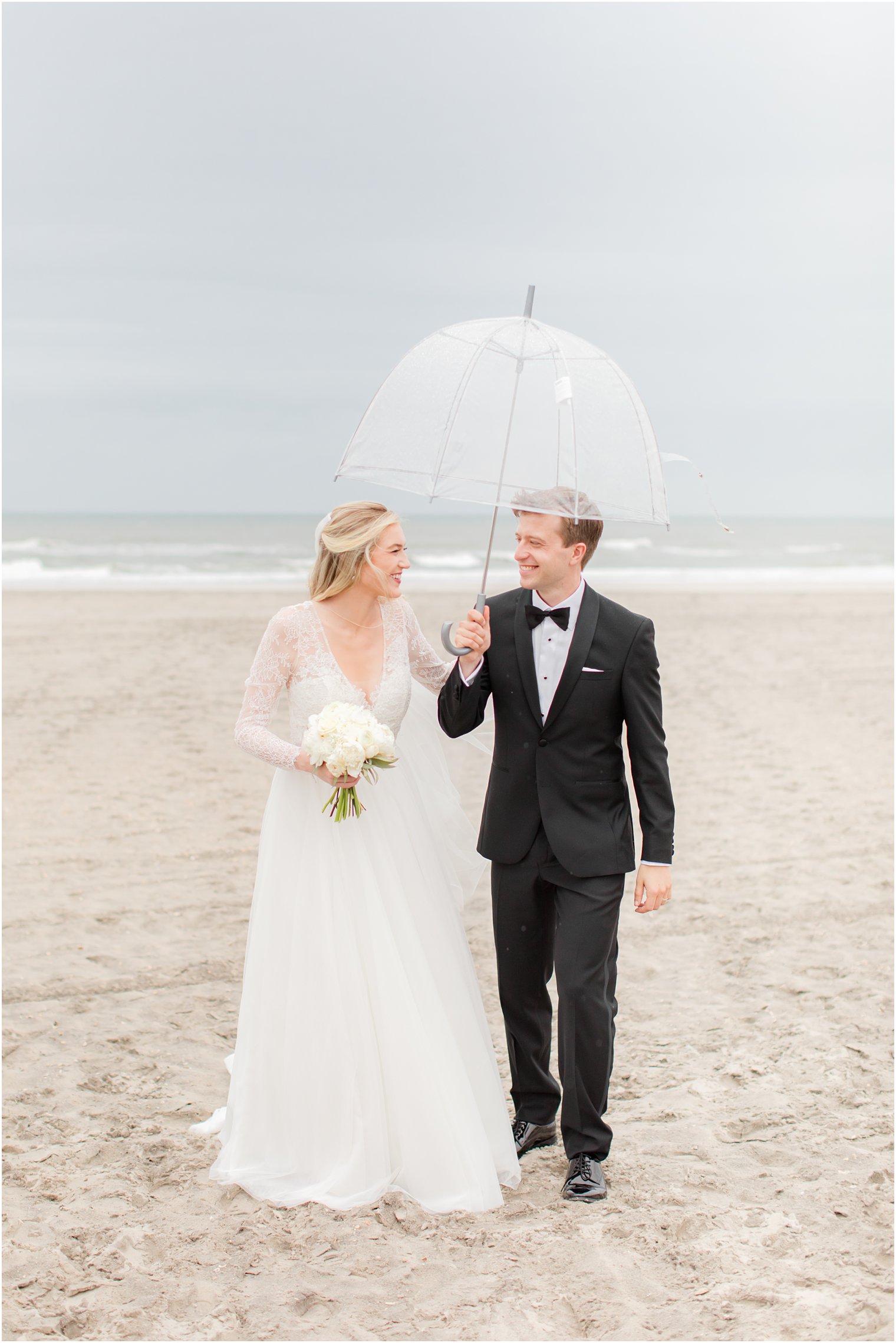 bride and groom hold clear umbrella walking on beach during wedding photos