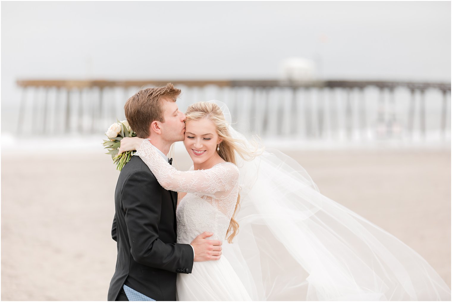 groom kisses bride's cheek during windy wedding portraits on Ocean City beach