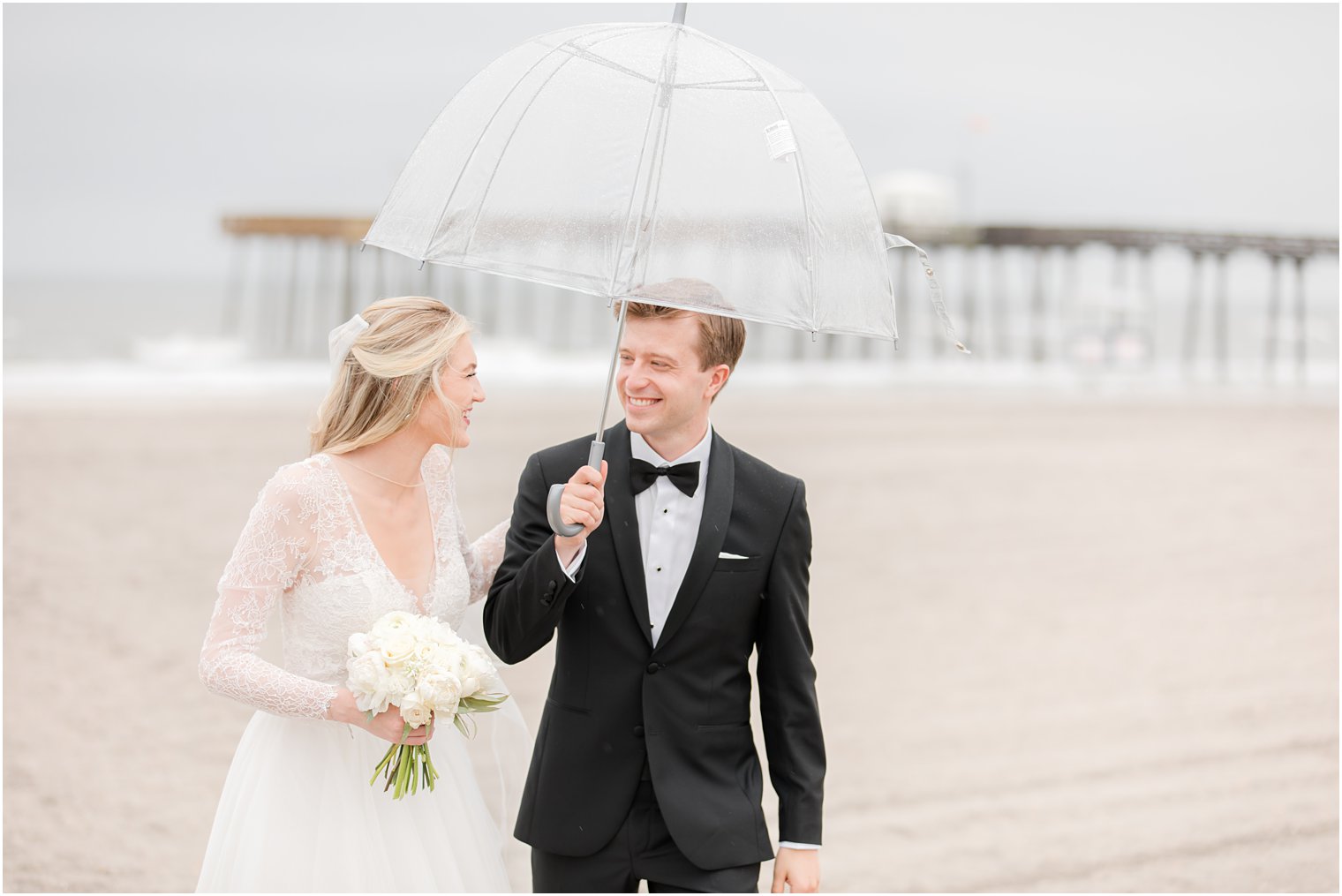 bride and groom walk on beach under clear umbrella on rainy wedding day