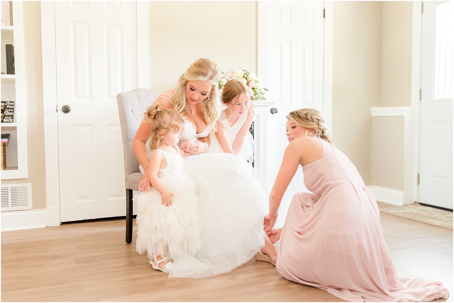 bride talks to flower girls while bridesmaid helps with shoes during wedding prep 