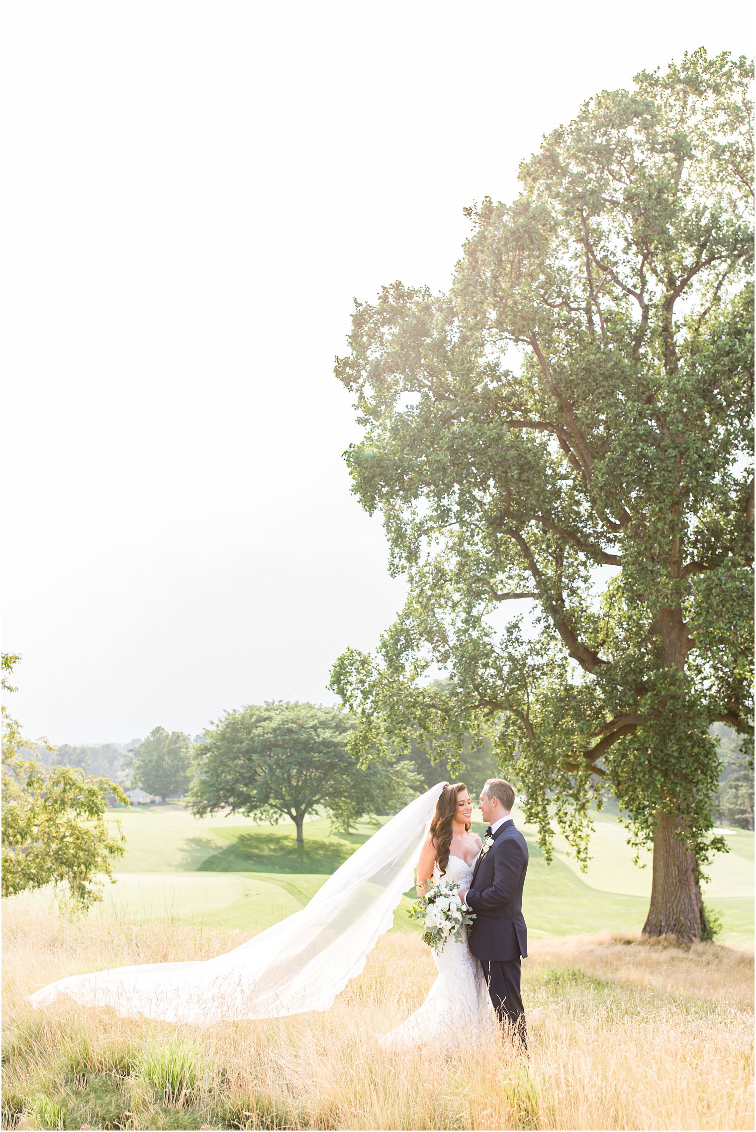 bride and groom pose in field with bride's veil behind her