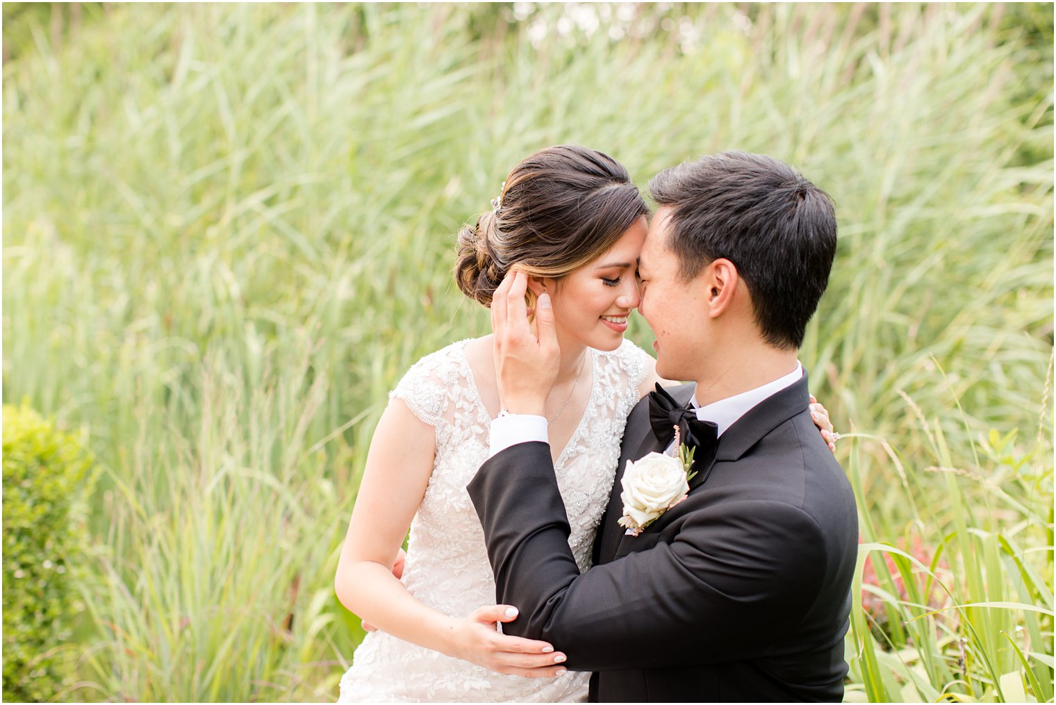 bride and groom sit in tall grass with foreheads touching 