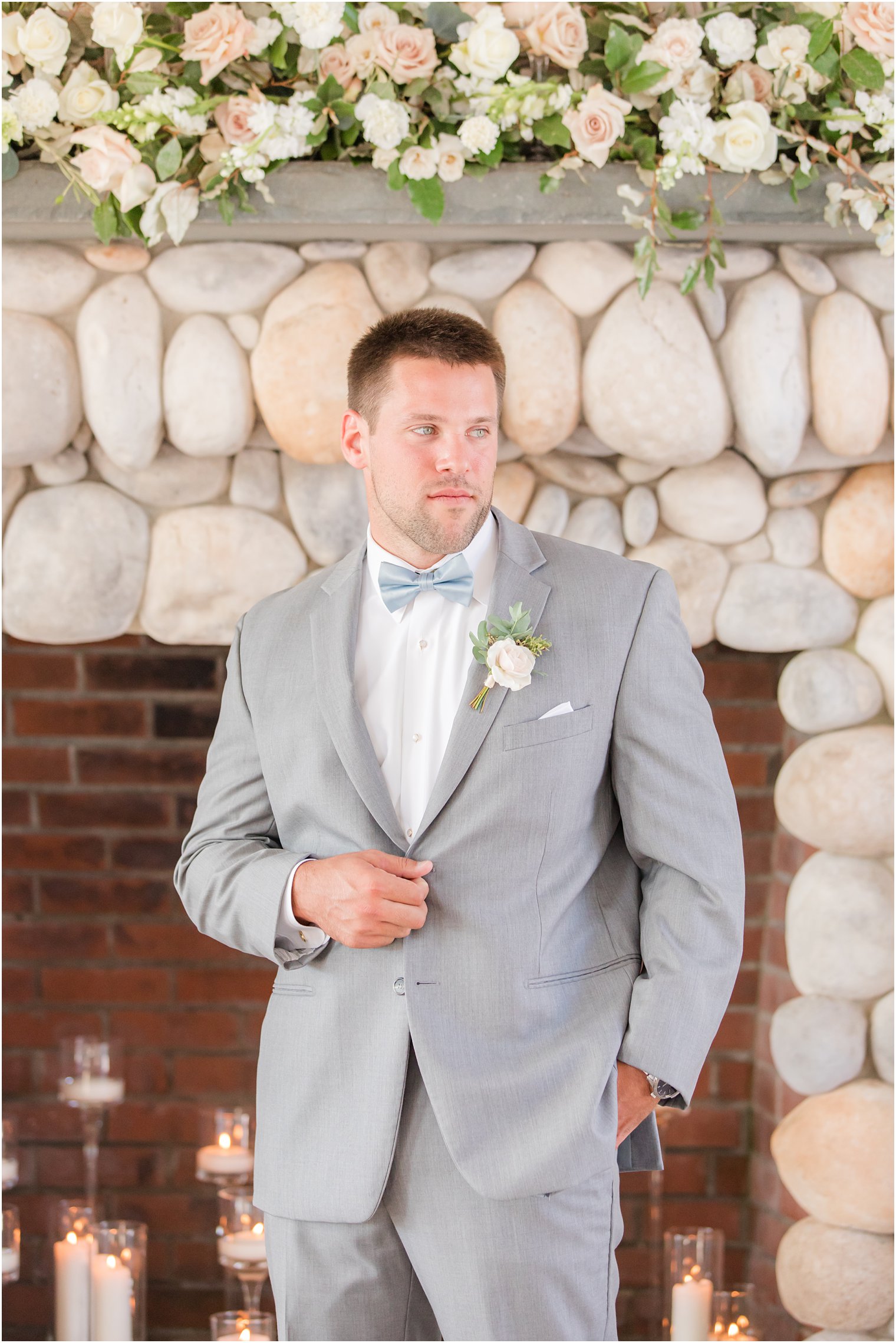 groom in grey suit poses in front of stone fireplace at Bonnet Island Estate