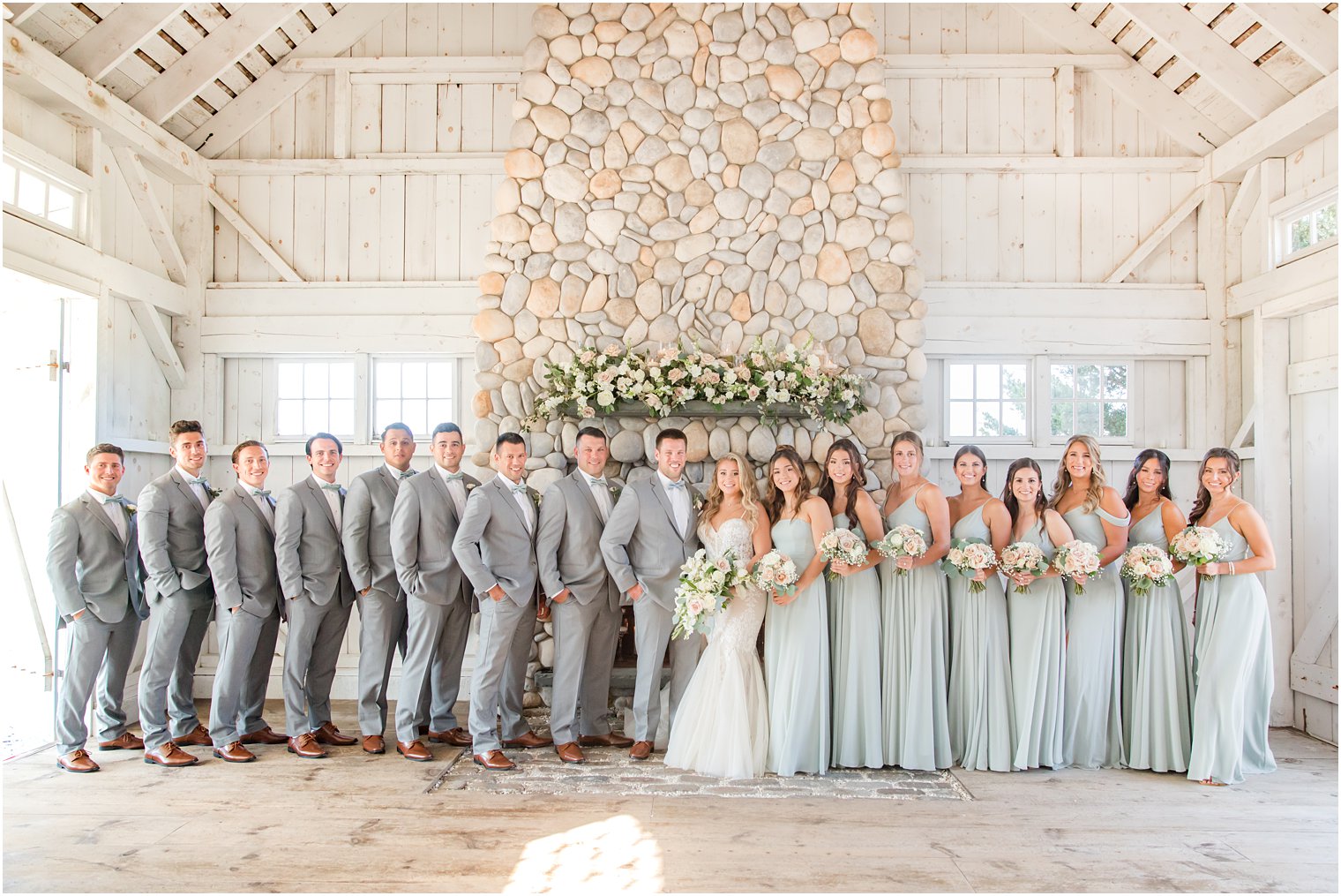 newlyweds pose with wedding party in grey suits and light blue gowns by stone fireplace