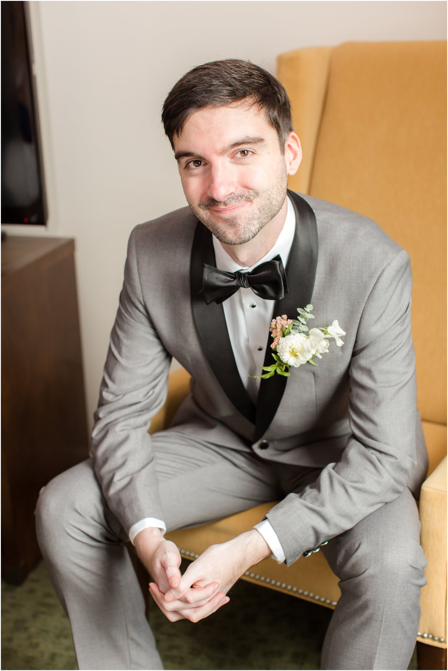 groom smiles sitting in chair in grey suit with black lapels