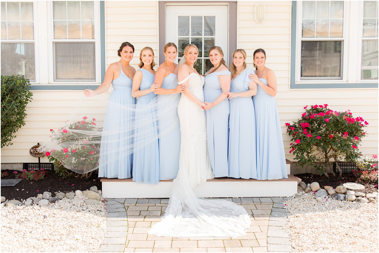 bride poses on steps of beach house with bridesmaids in light bleu gowns