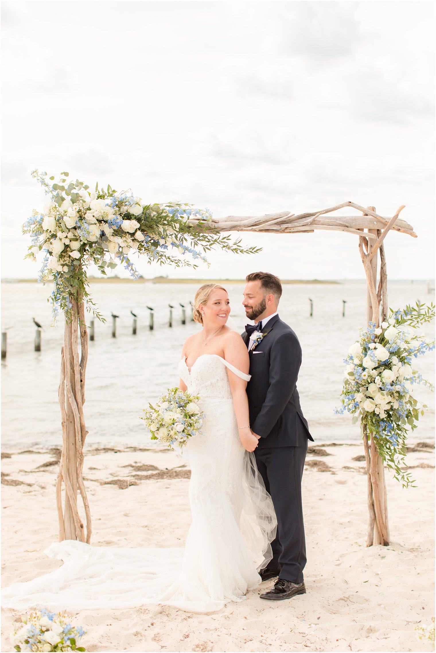 bride in off-the-shoulder gown poses with groom under wooden arbor with floral accents