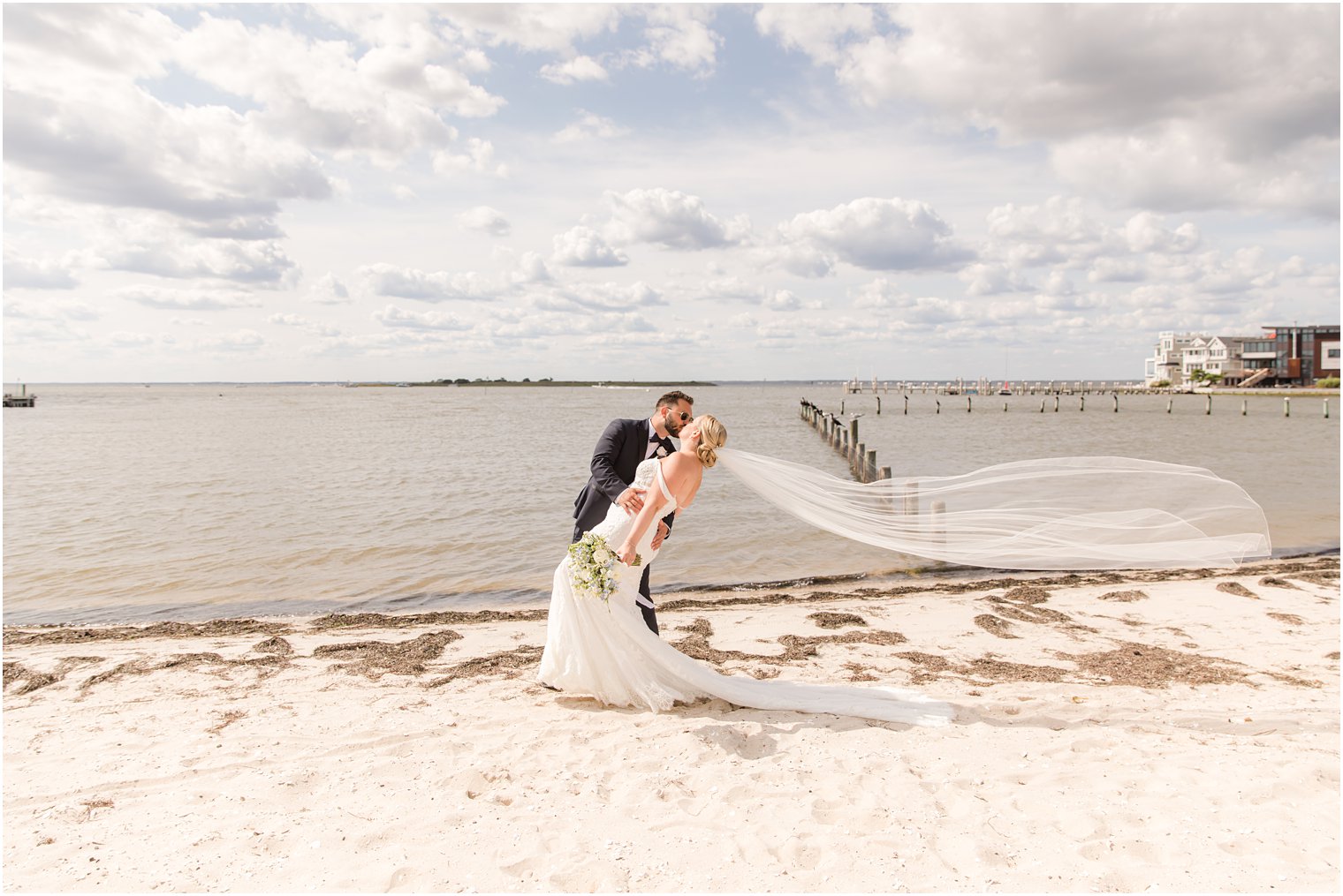 groom kisses bride dipping her on beach with veil floating behind them 