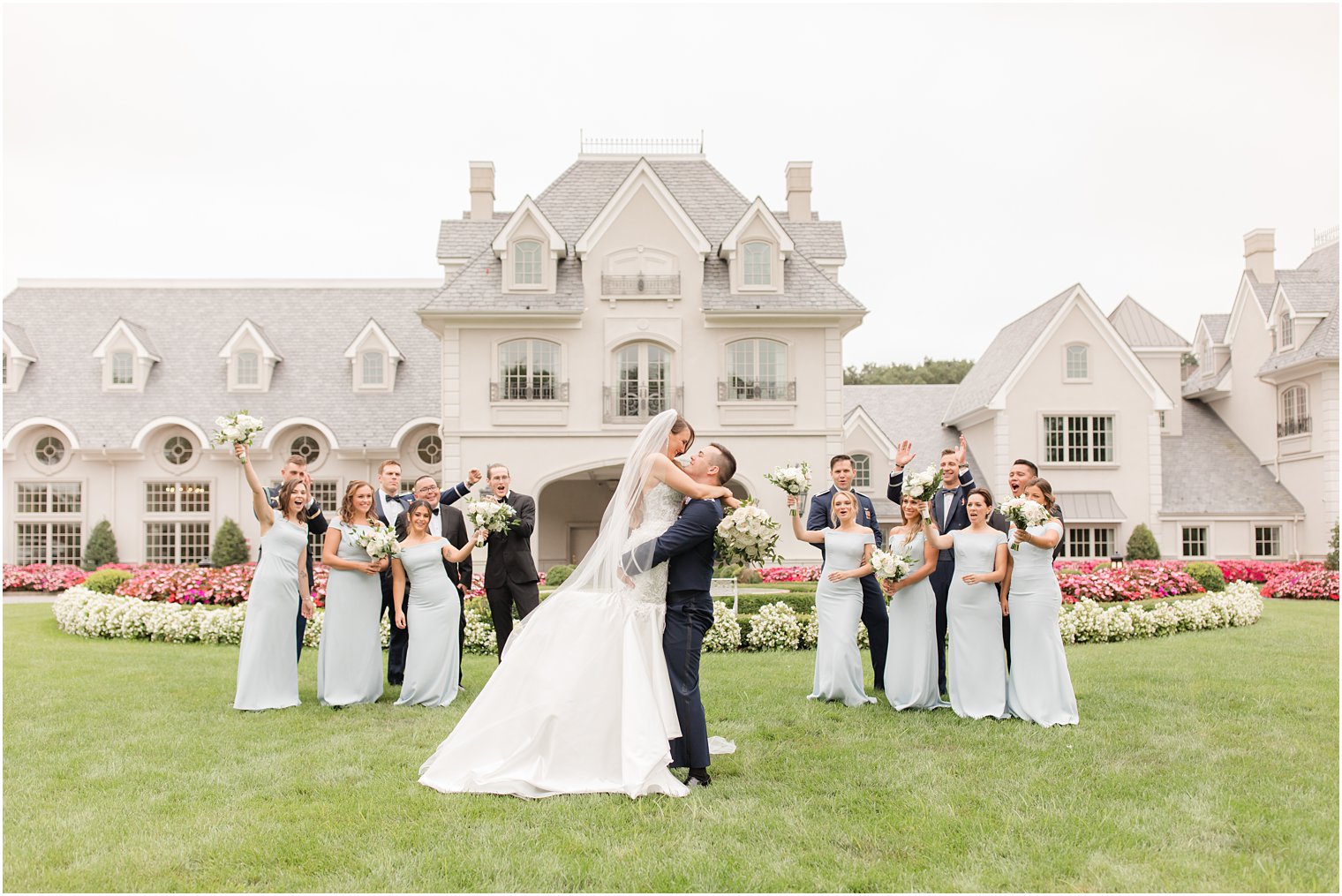 groom lifts bride up on lawn of Park Chateau Estate with wedding party behind them