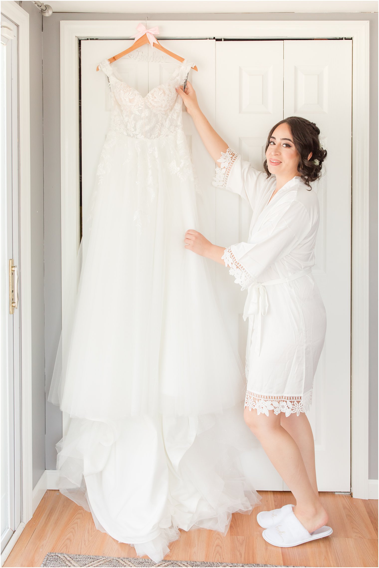 bride stands next to wedding dress in white robe and slippers 