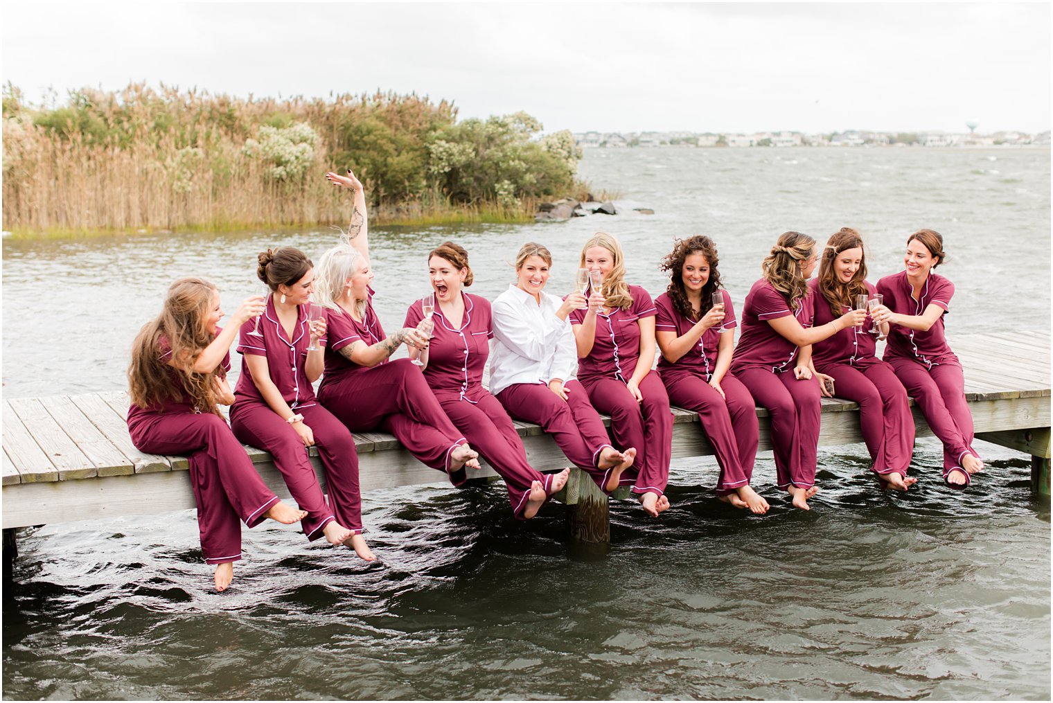 bridesmaids sit on dock at Bonnet Island Estate in matching burgundy pajamas 