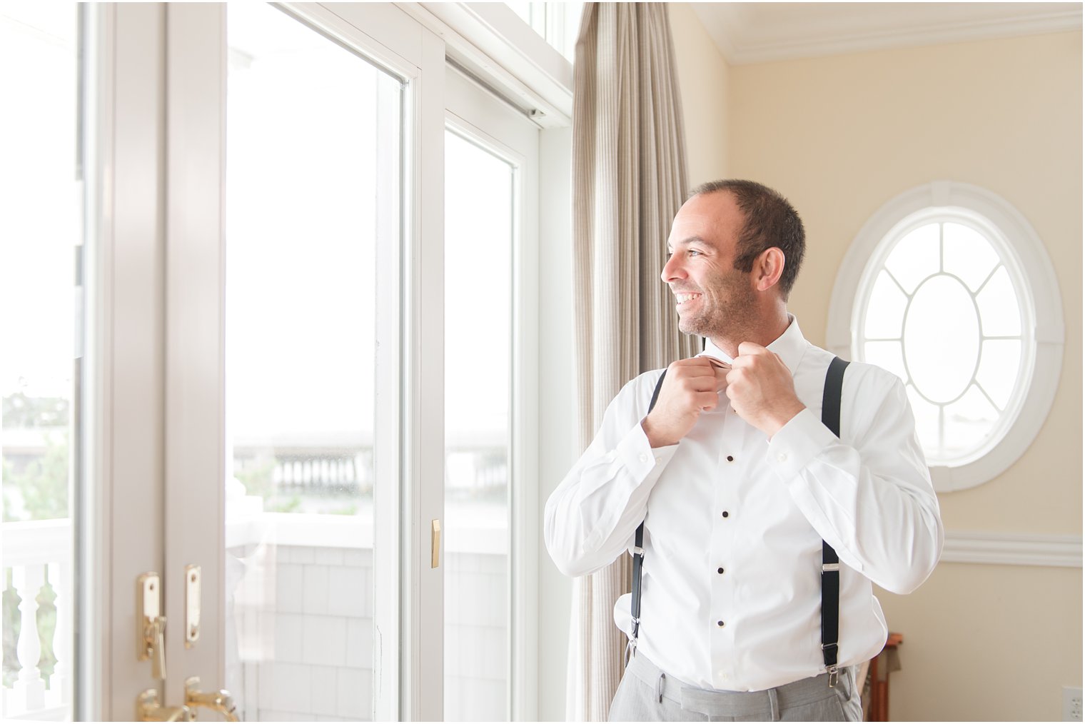 groom adjusts bowtie looking out window before wedding day