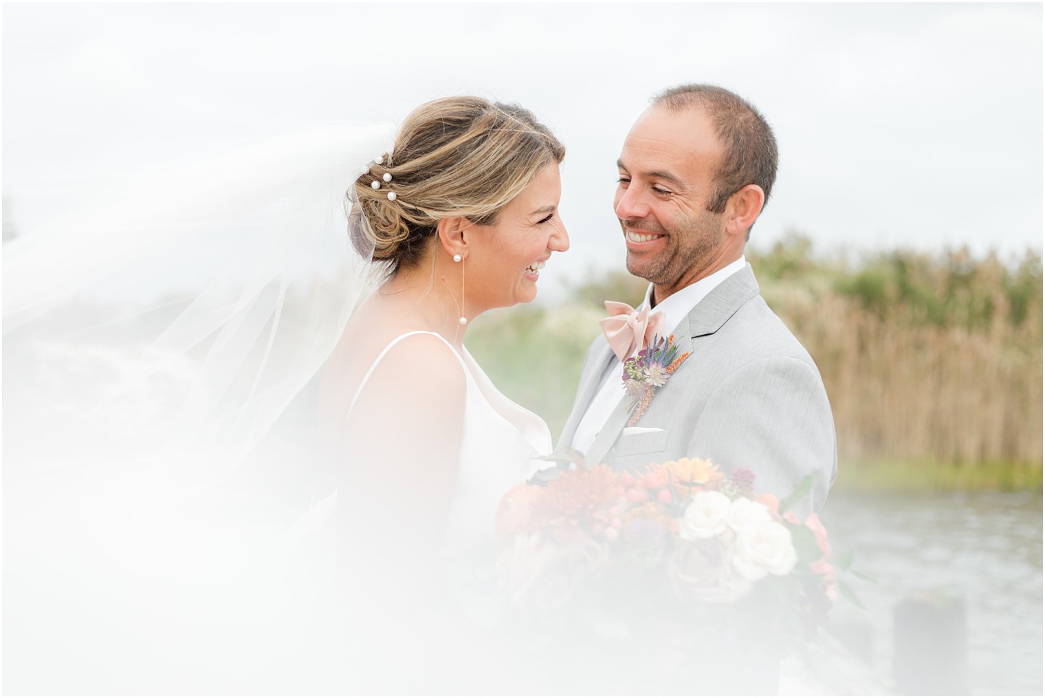 bride and groom laugh together as veil wraps around their shoulders 