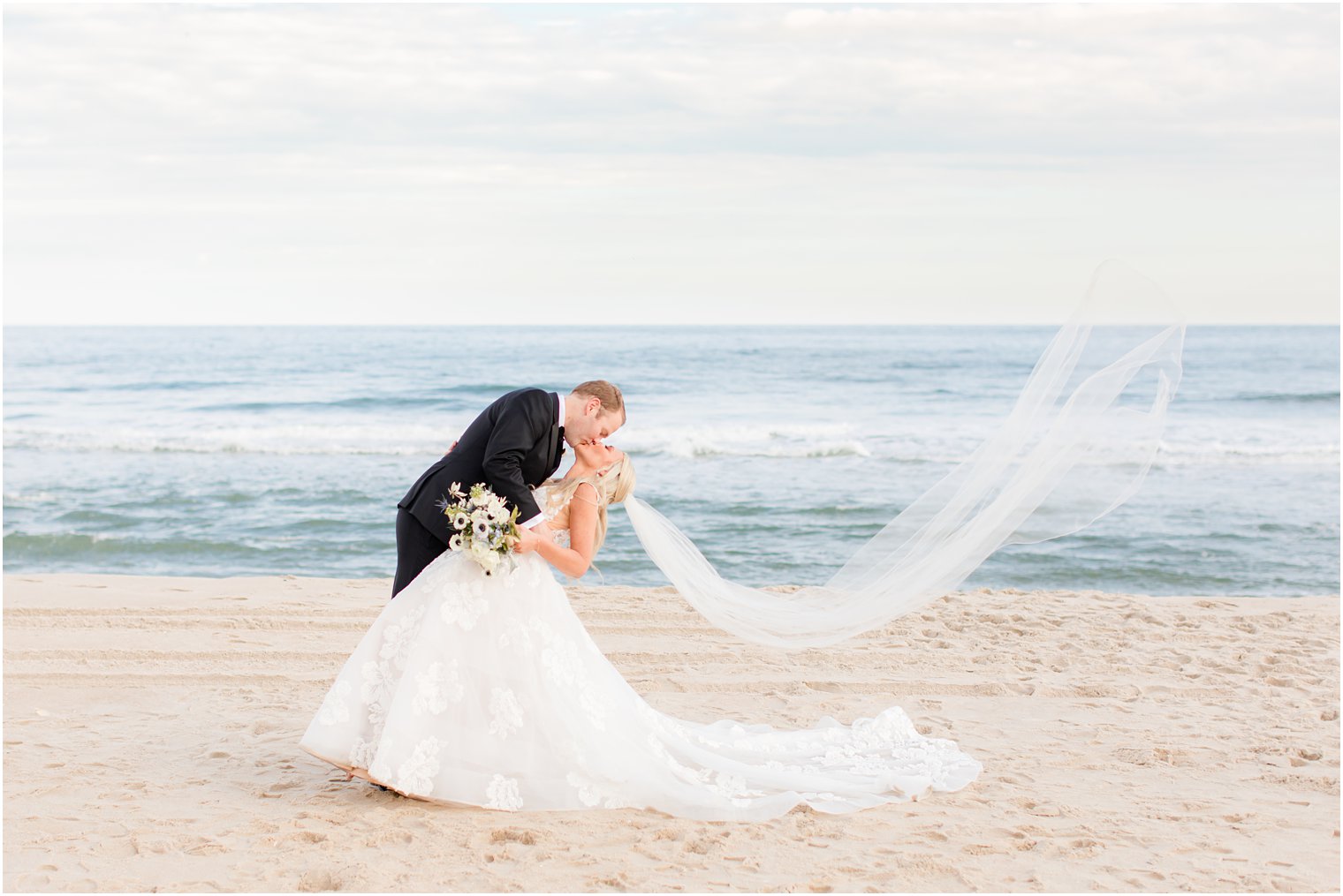 groom kisses bride dipping her while veil floats behind her on the beach 