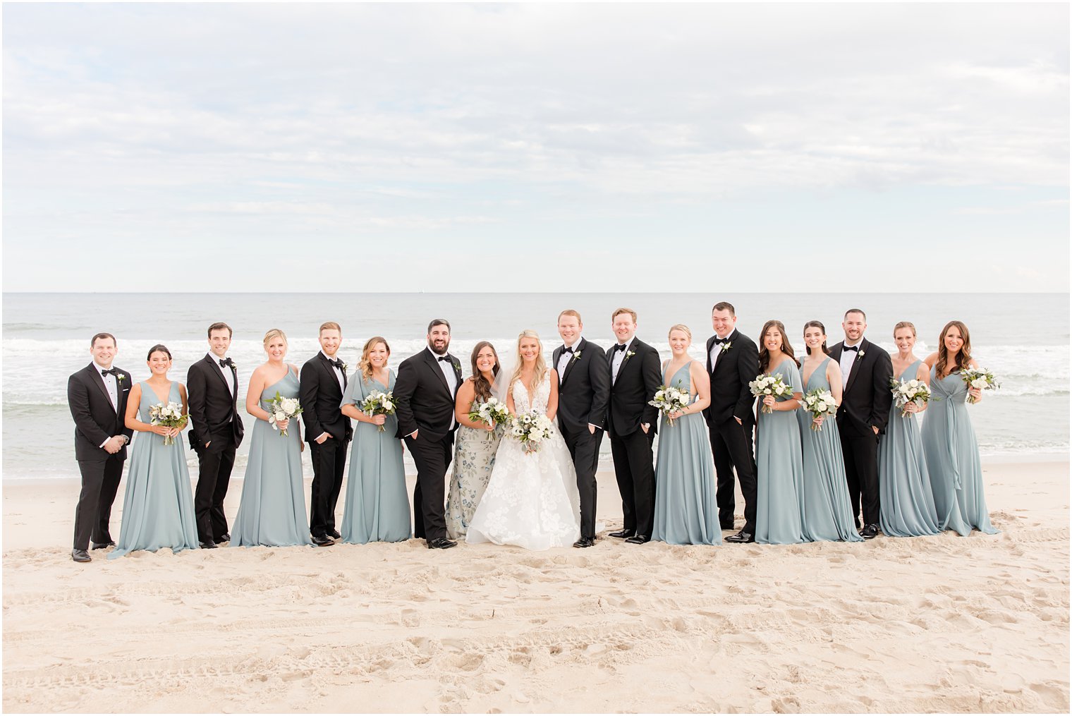 bride and groom stand with wedding party in black and blue on the beach 