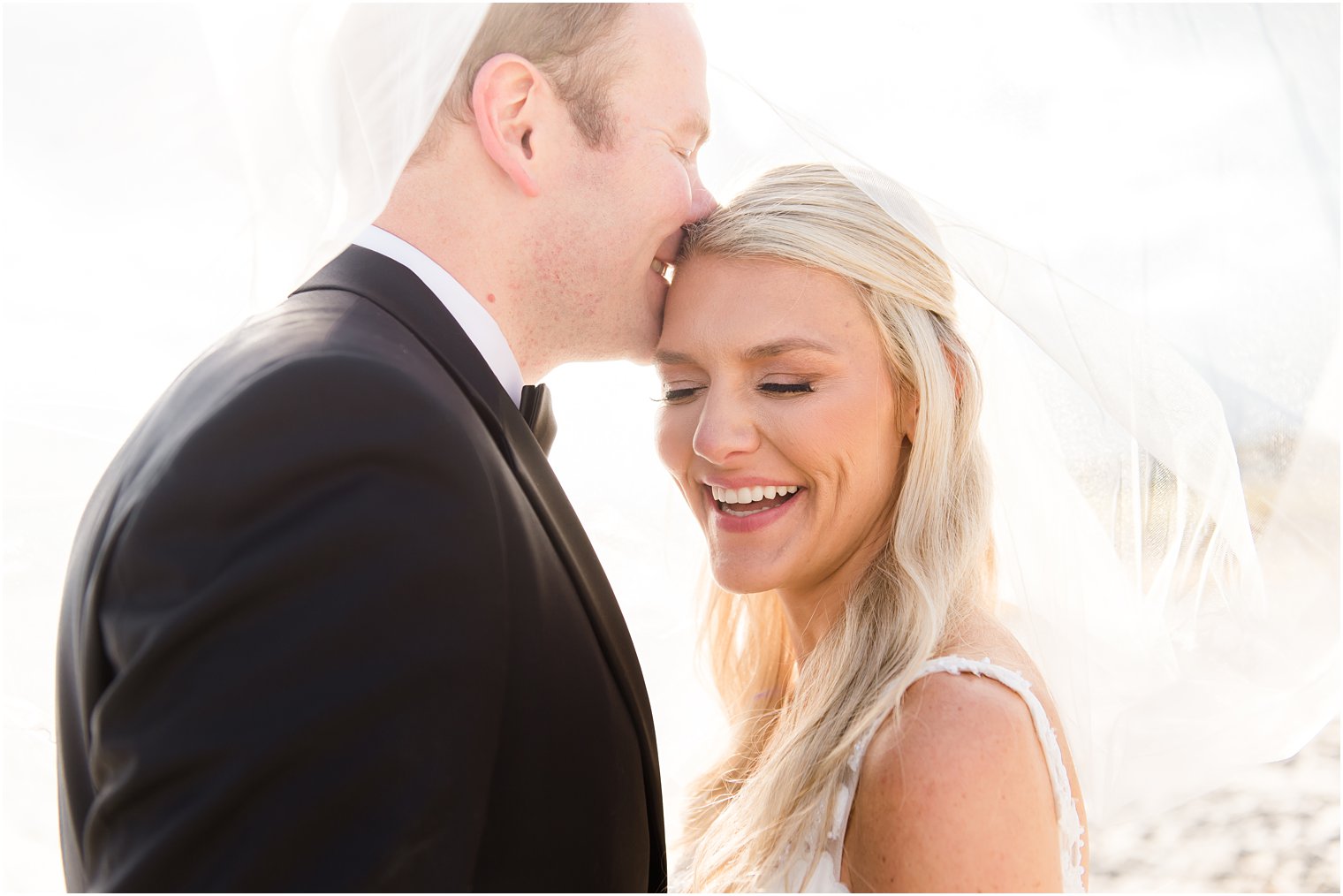 groom laughs nuzzling bride's forehead under her veil on the beach 