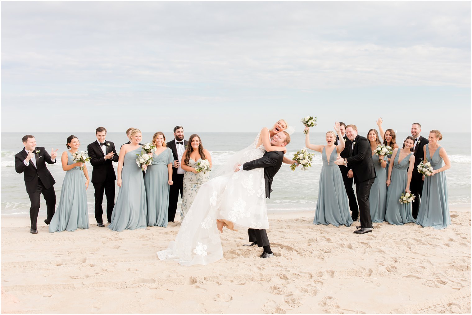 groom lifts bride laughing while bridal party cheers on beach 