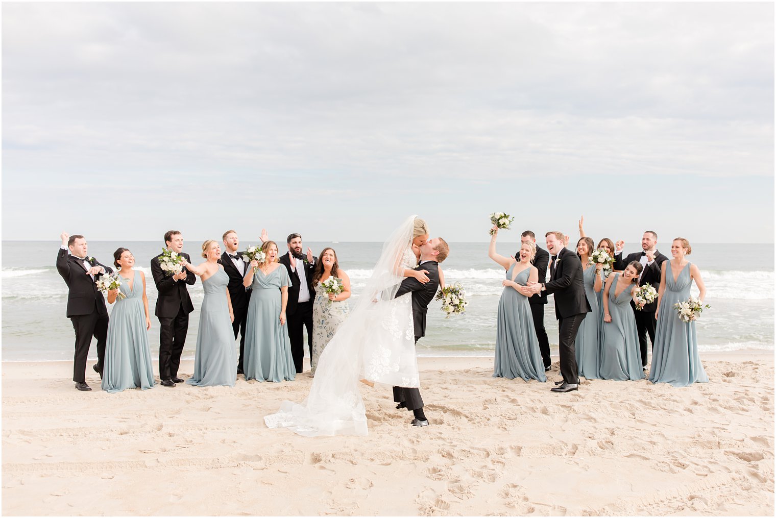 wedding party cheers as groom lifts up bride on beach kissing her 