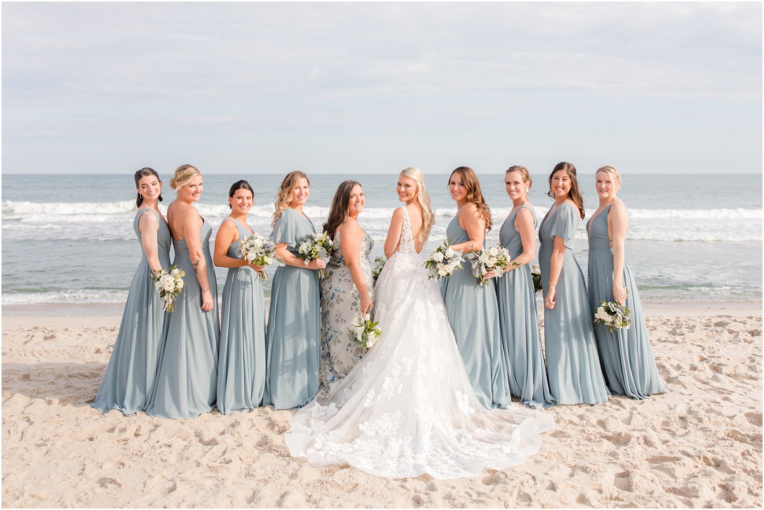 bride smiles looking over shoulder with bridesmaids in light blue gowns on the beach 