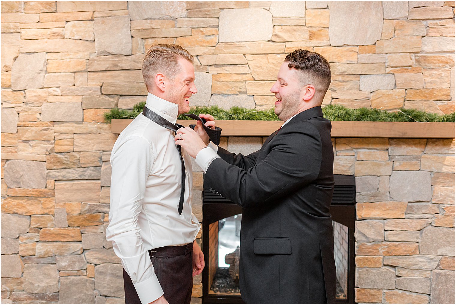 groomsman helps groom with tie in front of stone fireplace at Bear Brook Valley