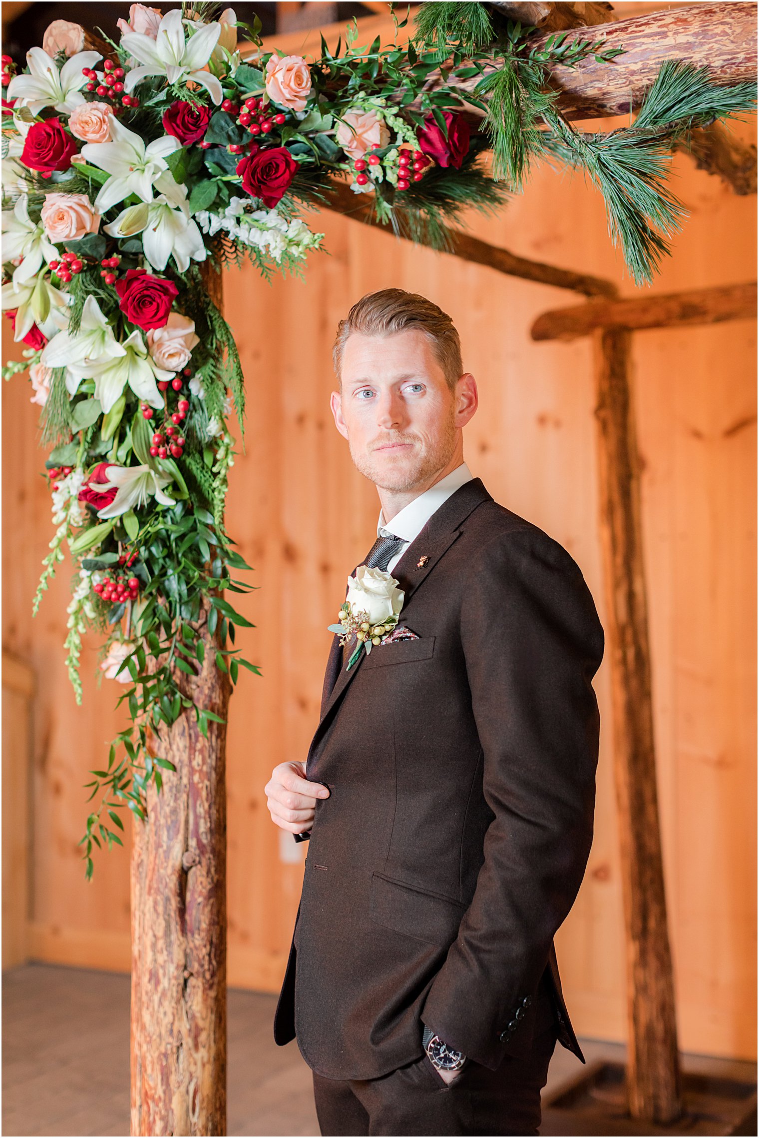 groom stands under wooden arbor with red and white flowers 