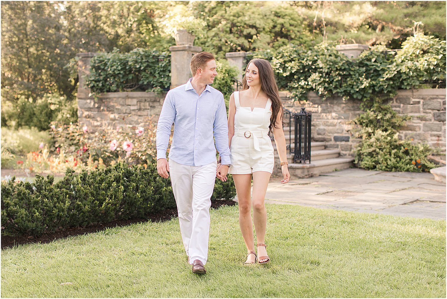 bride and groom hold hands walking through garden by stone wall