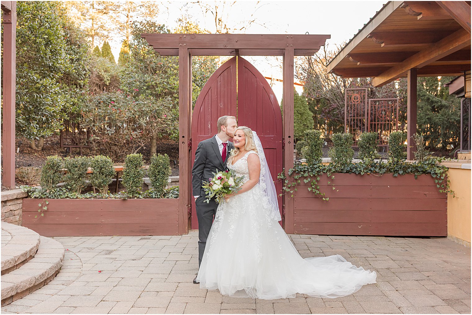 groom kisses bride's cheek by red gate