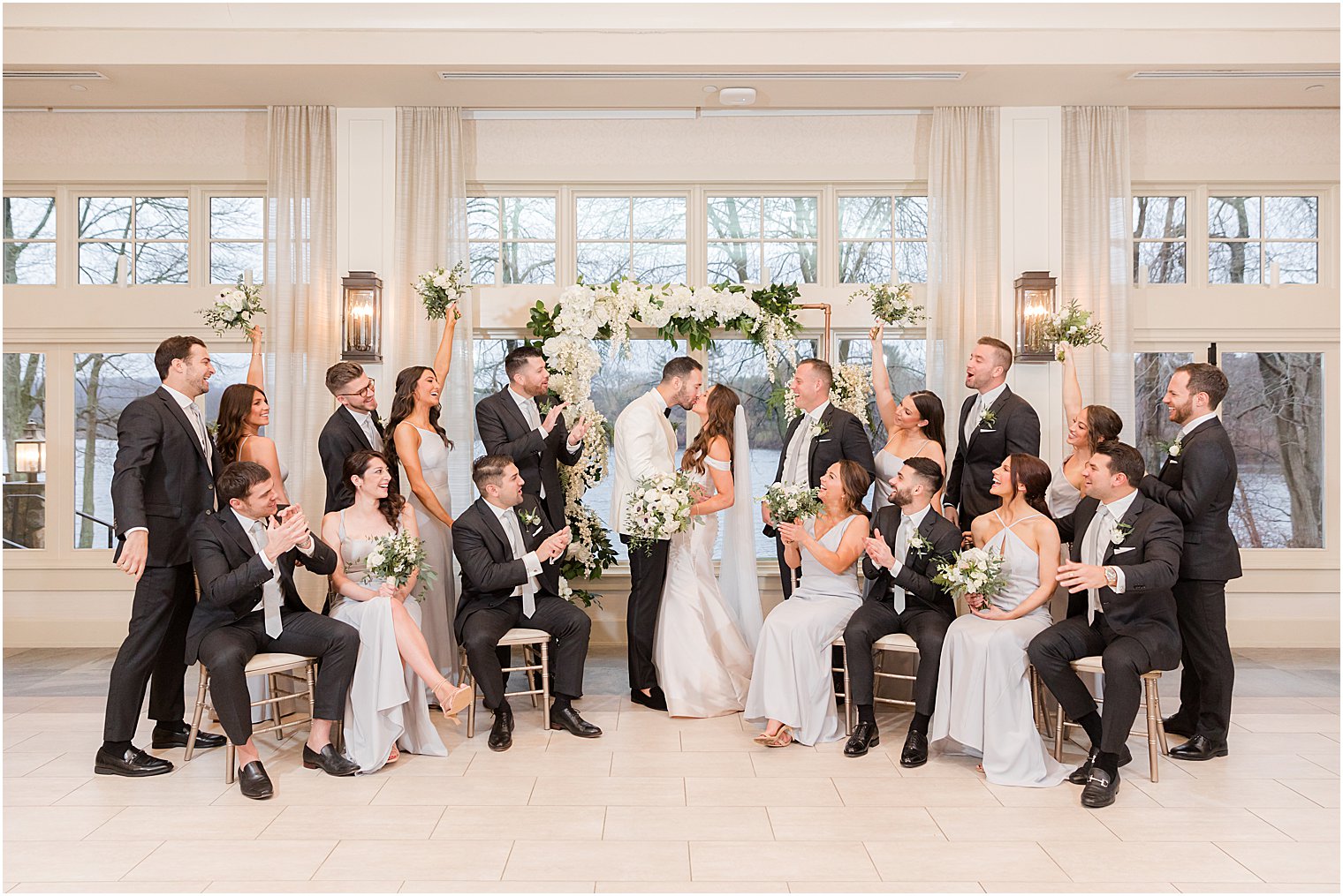 bride and groom kiss by floral arch and bridal party surrounds them