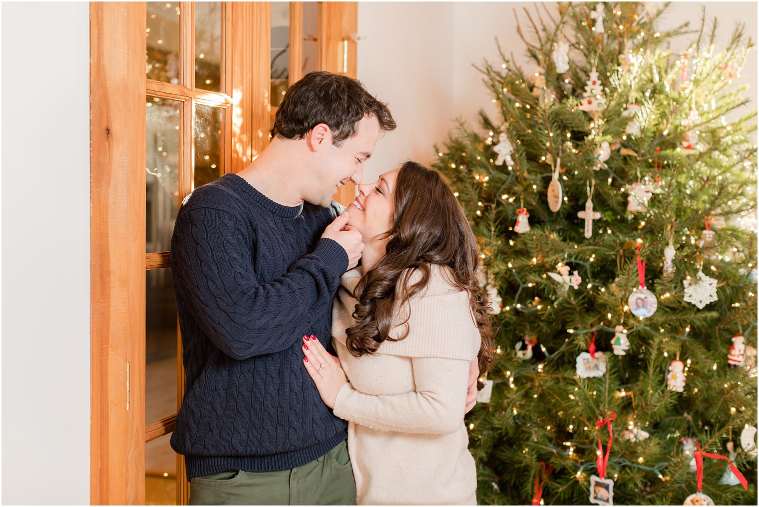 groom lifts bride's chin to kiss her by Christmas tree during at home engagement session 
