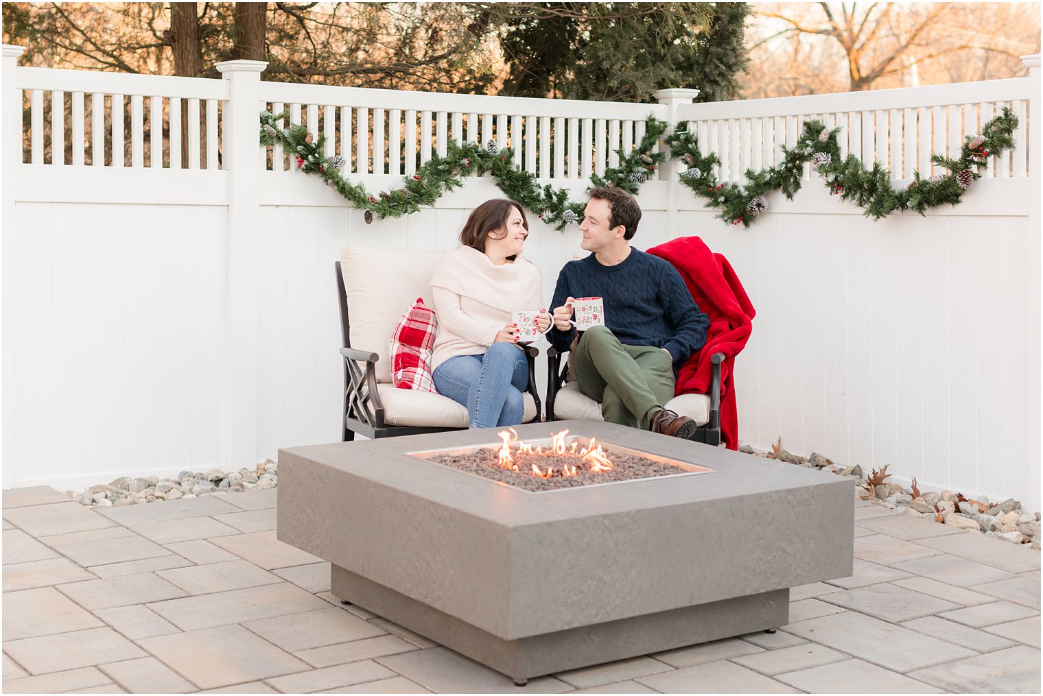 engaged couple sits by fire pit holding holiday mugs during engagement photos at home 
