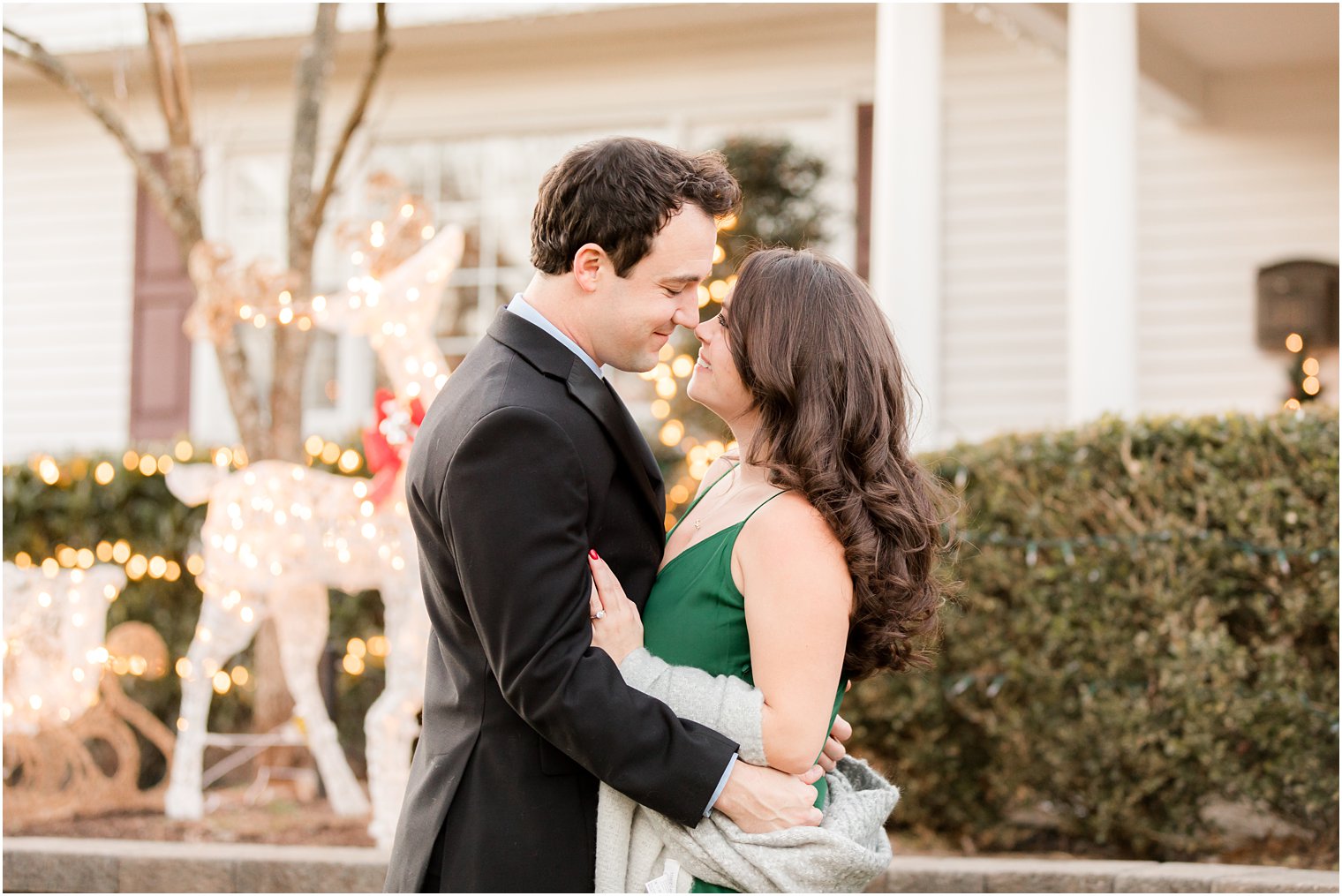 bride and groom stand with noses touching on front lawn during holiday engagement session 